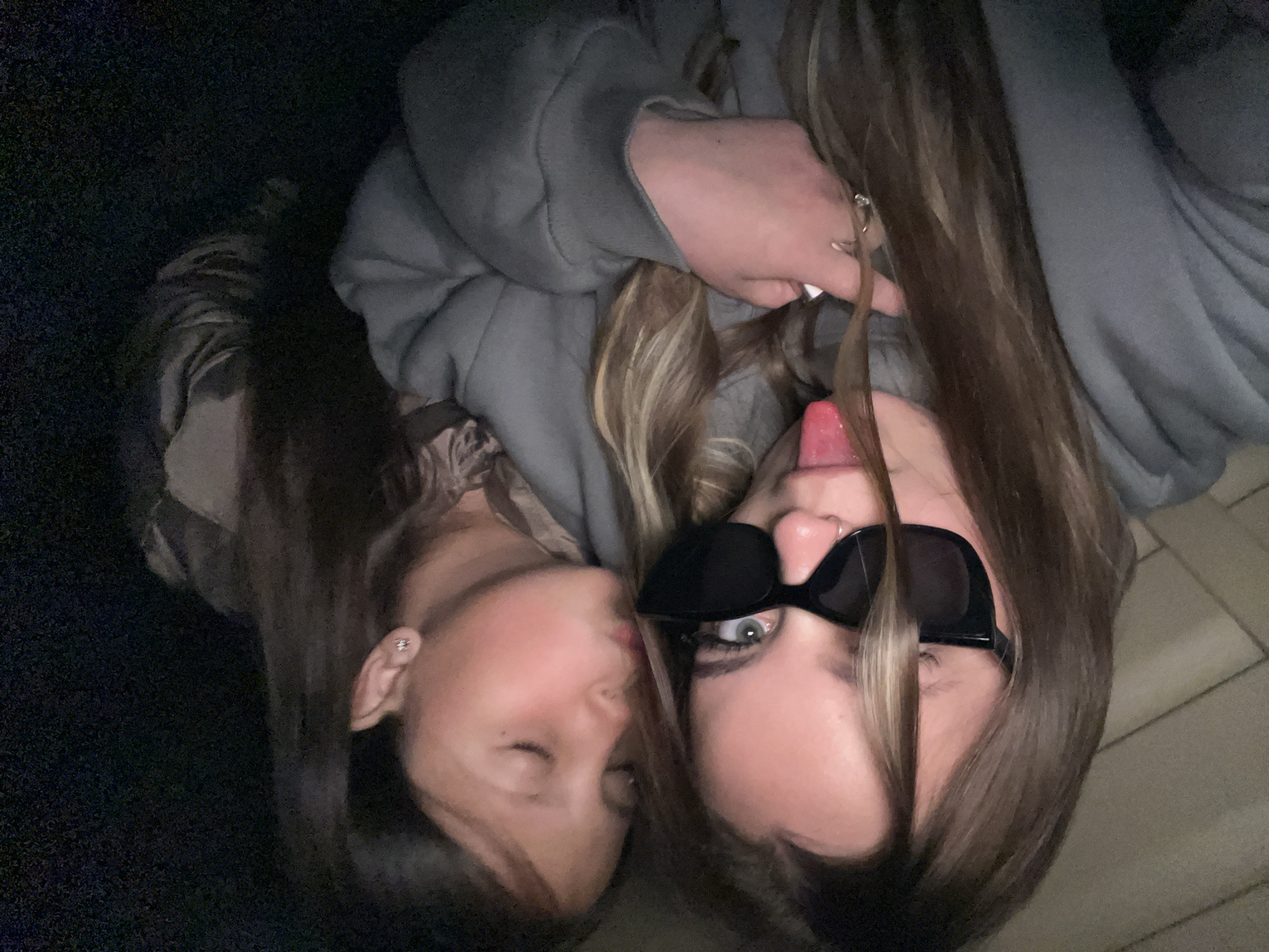Two women are shown in a close-up photograph. One woman is wearing sunglasses and sticking out her tongue while holding a strand of hair. The other woman has her eyes closed. Both are wearing casual clothing. The location is not discernible from the image.