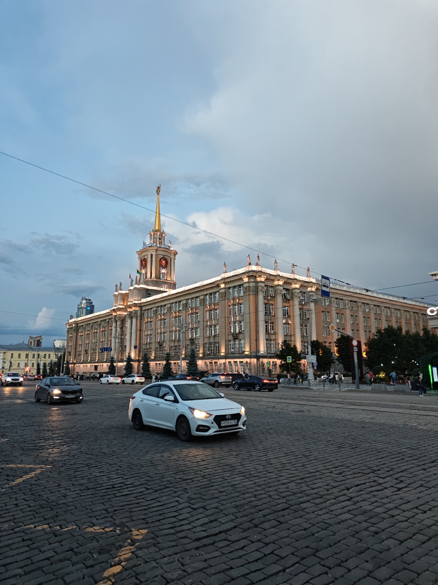 The image captures a wide shot of the Yekaterinburg City Administration building, also known as the "House of Soviets," located at 1 Square of 1905, Yekaterinburg, Russia. The building is illuminated and features a prominent golden spire with a clock tower. The sky is overcast with a mix of blue and gray clouds. In the foreground, a cobblestone street is visible with several cars driving, including a white Hyundai sedan in the immediate foreground. Pedestrians are present on the sidewalk in front of the building, and trees line the facade. The overall scene suggests an urban environment during the late afternoon or early evening.