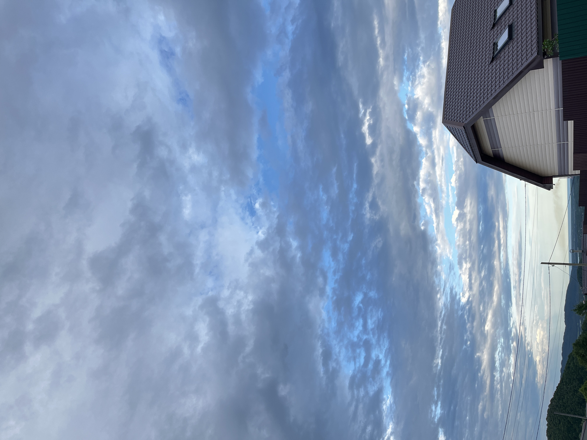The image shows a view of the sky and a house. The sky is filled with clouds, some of which are dark gray and heavy, while others are lighter and allow patches of blue sky to show through. There are some scattered white clouds, particularly near the horizon.
In the foreground, the roof and upper part of a house are visible. The roof is dark brown and has a steep pitch. There are two skylights on the roof. The siding of the house is light beige with horizontal stripes of gray. A dark brown fence is also visible in front of the house.
In the distance, there are rolling hills covered in green trees. Power lines stretch across the scene, and a utility pole is visible on the right side of the image. The overall atmosphere is overcast, with the light suggesting it could be late afternoon or early evening. The scene is a residential area, likely in or near a rural or suburban part of Petropavlovsk-Kamchatsky, Russia, given the architectural style and the presence of natural landscape elements like hills and trees. There are no people or animals visible in the image.
