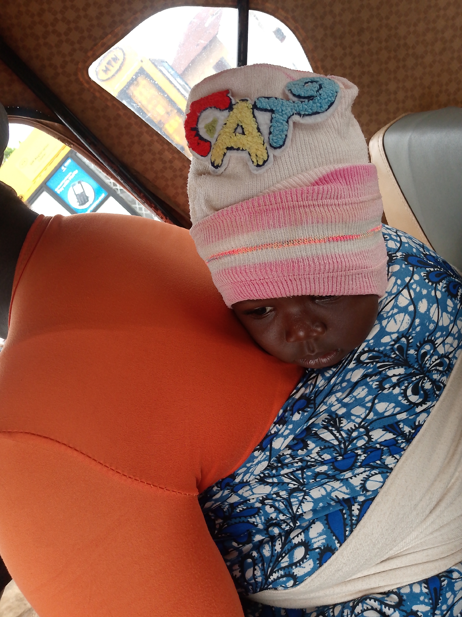 A child is being held inside a vehicle. The child is wearing a hat with the name "CATE" on it. The vehicle appears to be a tuk-tuk, as evidenced by the interior roof pattern and window. The location seems to be somewhere with MTN advertising.