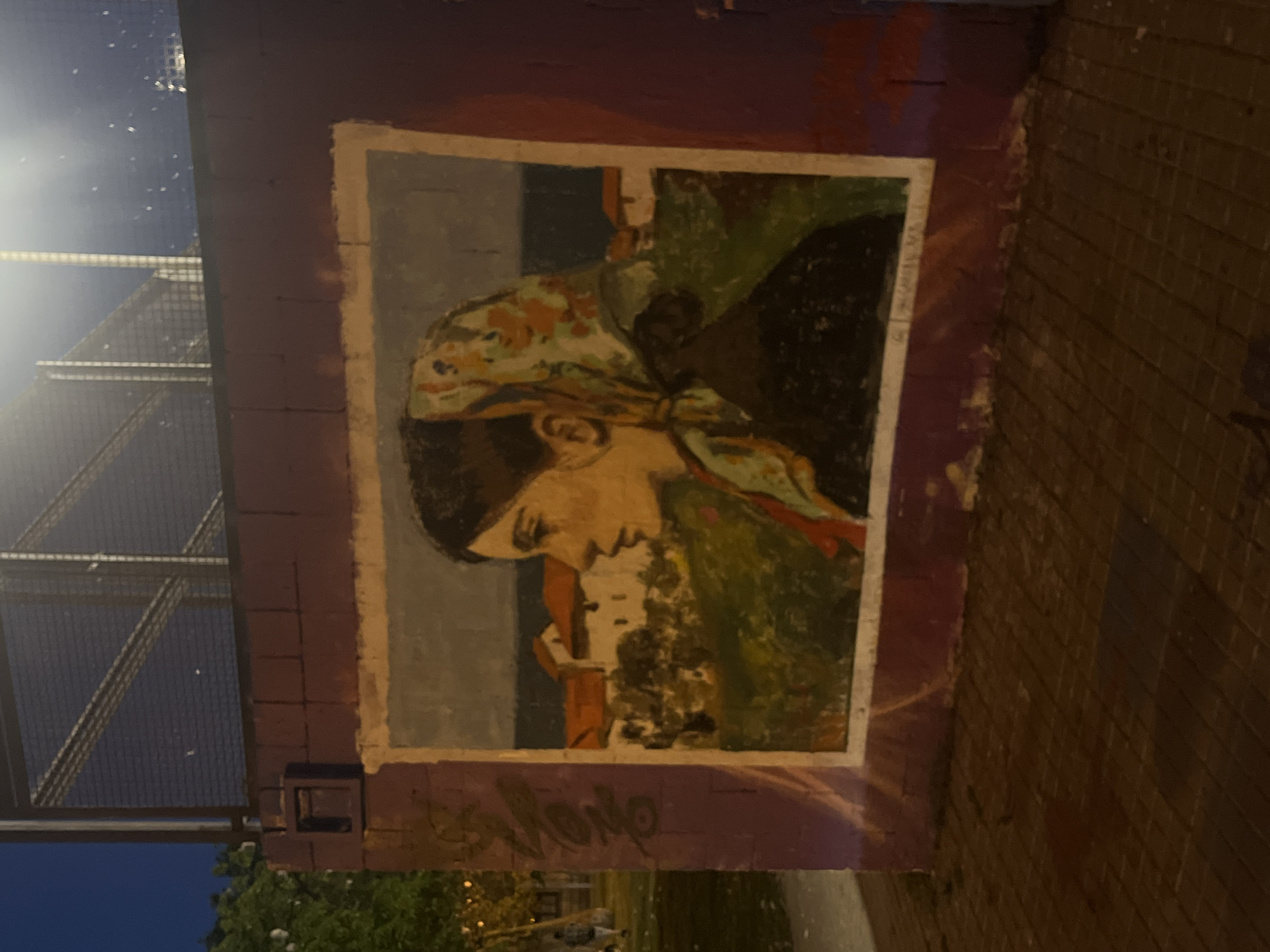 The image shows a painted mural on a brick wall, likely outdoors. The mural depicts a woman in profile wearing a headscarf, set against a landscape with houses or buildings in the background. The painting is framed by a light border and the surrounding wall is painted a dark reddish-purple. The mural is also vandalized by graffiti. There is a metal structure visible to the left of the mural, suggesting an urban environment. Based on the style of the mural and the urban environment, it is possible that this mural is located in Barcelona, Spain, although confirmation would require additional information or visual cues. The scene appears to be photographed at dusk or night, given the presence of a light source and the darkness of the surrounding environment.