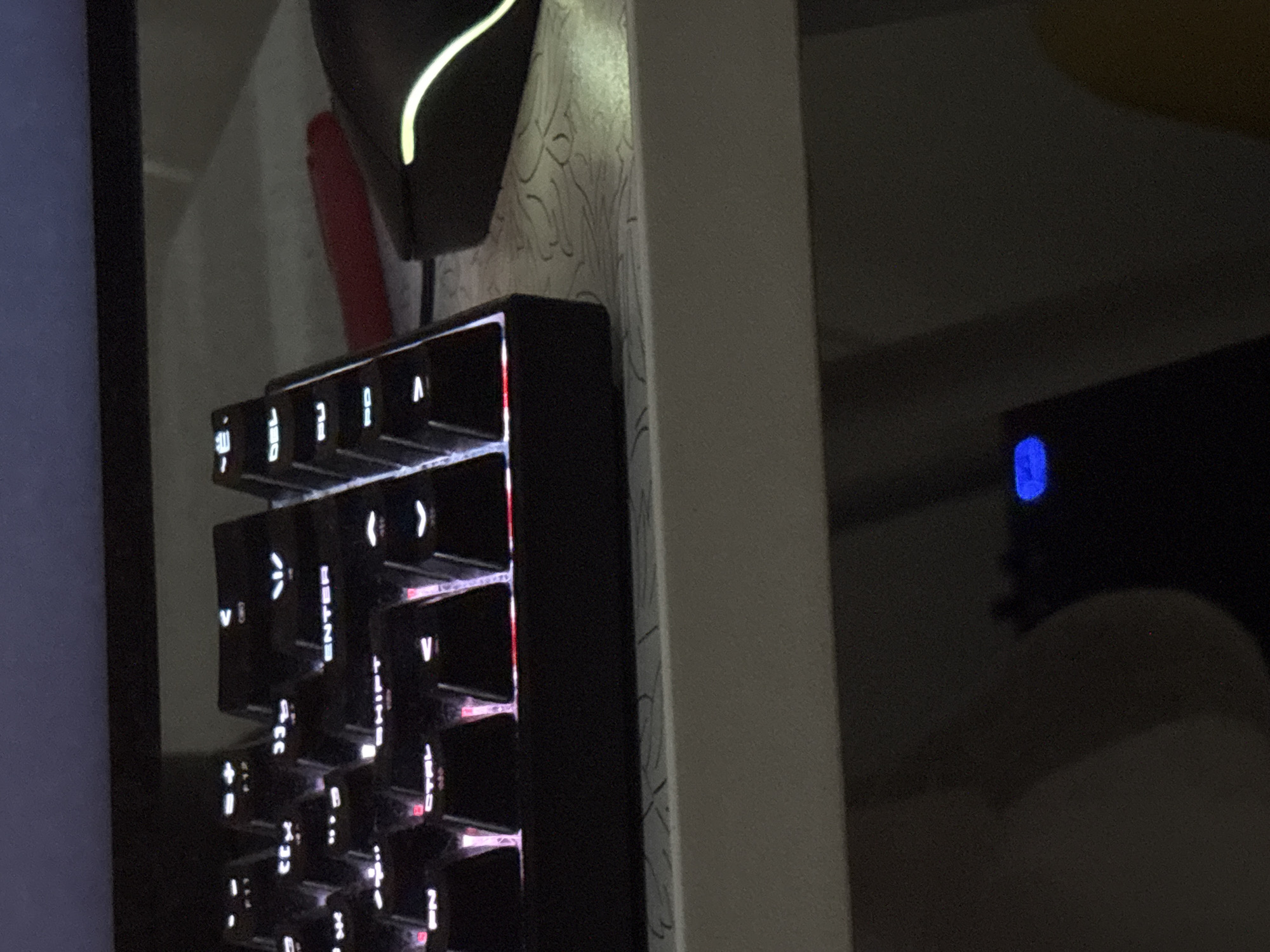 A close-up shot depicts a black, vertically-oriented keyboard with illuminated white lettering and red backlighting. The keyboard is positioned next to a white surface with a patterned wallpaper. A black gaming headset with a glowing white accent line rests above the keyboard. In the blurred background, a dark monitor displays a blue light. The specific location in the city of Chaikovsky, Russia, cannot be determined from the available information.