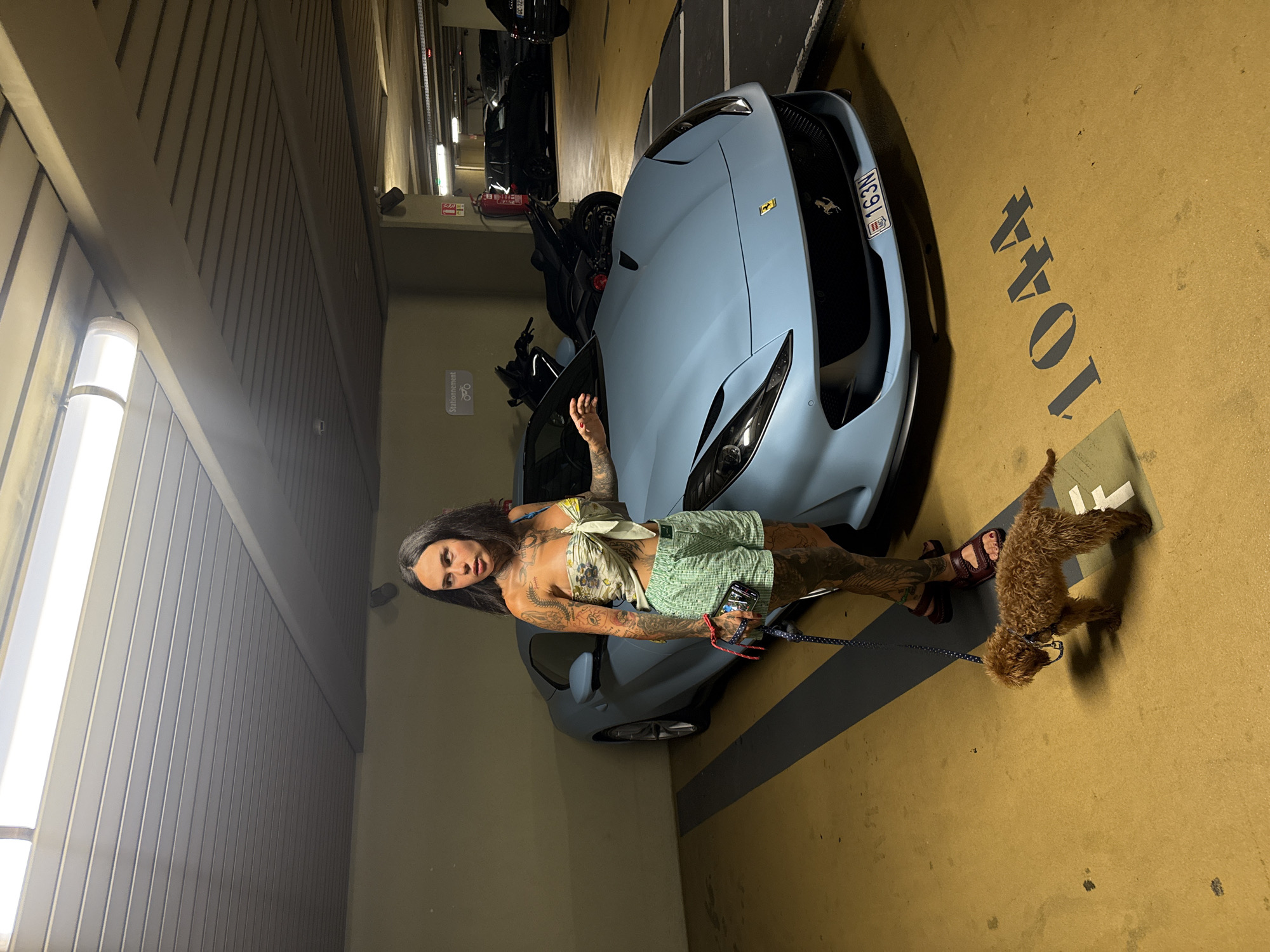 A woman with extensive tattoos stands next to a light blue sports car in a parking garage. She is wearing a green and white checkered skirt and a white halter top. A small, fluffy brown dog is on a leash, standing near her feet. The car has a license plate that reads "MC 163N". The parking spot is marked with the number "1044". The scene appears to be indoors, likely in a parking garage, with artificial lighting. The overall impression is one of a casual outing with a pet and a luxury vehicle.