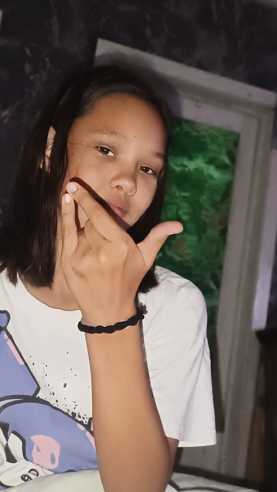 The media file shows a close-up, head-and-shoulders shot of a young person with dark hair and brown eyes. The person is wearing a white t-shirt with a colorful graphic on the left side. They are holding their right hand up, with their index and pinky fingers extended in a gesture commonly known as the "rock on" or "devil horns" sign. A black braided bracelet with white trim is visible on their wrist.
The background is dimly lit and appears to be an indoor setting. To the left, there is a dark, textured surface, possibly a wall. To the right, a window or doorway is visible, with a blurred green curtain or material behind it. The lighting suggests it could be nighttime or a dimly lit room, and there are no clear indicators of weather or specific location cues beyond being indoors. The person's expression is neutral to slightly playful. There is no visible text in the image. The location context provided is Karsha, Russia.