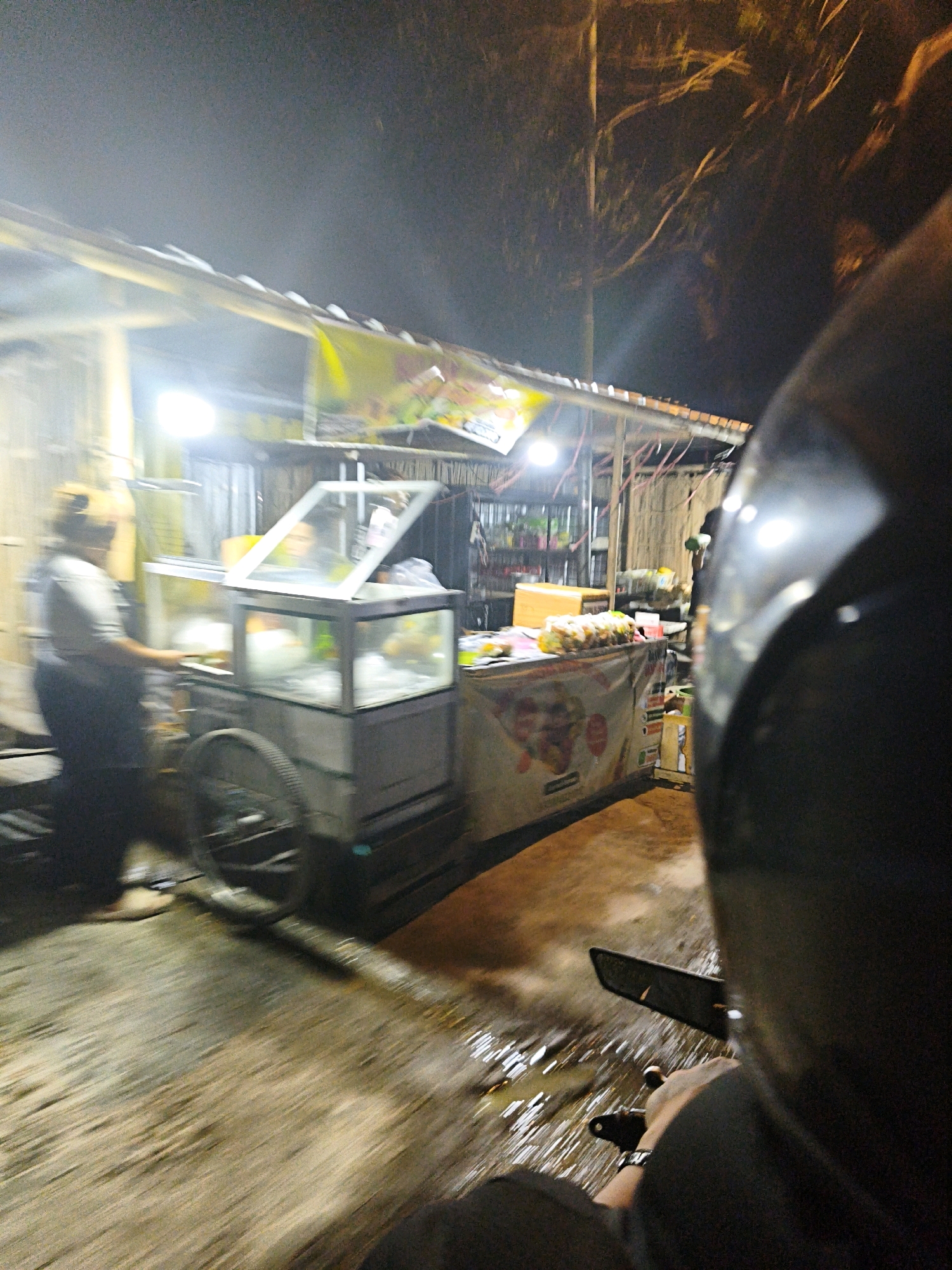 This is a low-light, motion-blurred image taken at night, likely from the perspective of a person riding a motorcycle, looking towards a street-side food stall in Desa Donohudan, Indonesia. The food stall appears to be a mobile cart with a glass display case, illuminated by overhead lights. A person, possibly a vendor, is partially visible to the left, standing near the cart. The cart has a large wheel on the left side. The ground in front of the stall is wet, reflecting the lights. In the foreground, a portion of a dark helmet and a hand holding a handlebar are visible, indicating the viewpoint of someone on a motorcycle. Trees and branches are visible against the dark night sky in the background, suggesting an outdoor, possibly rural or suburban, setting within the village.