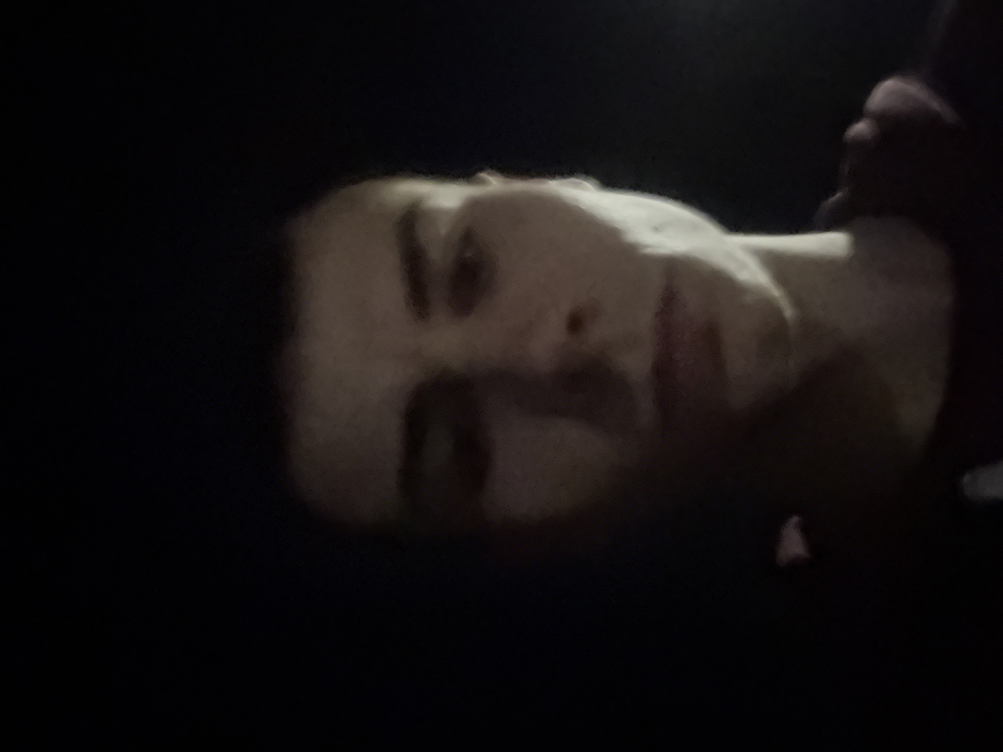 A low-angle, blurry, and dark photograph of a person's face, likely taken in low-light conditions. The person has dark hair and appears to be male. The background is almost entirely black. There is no information to discern the location.