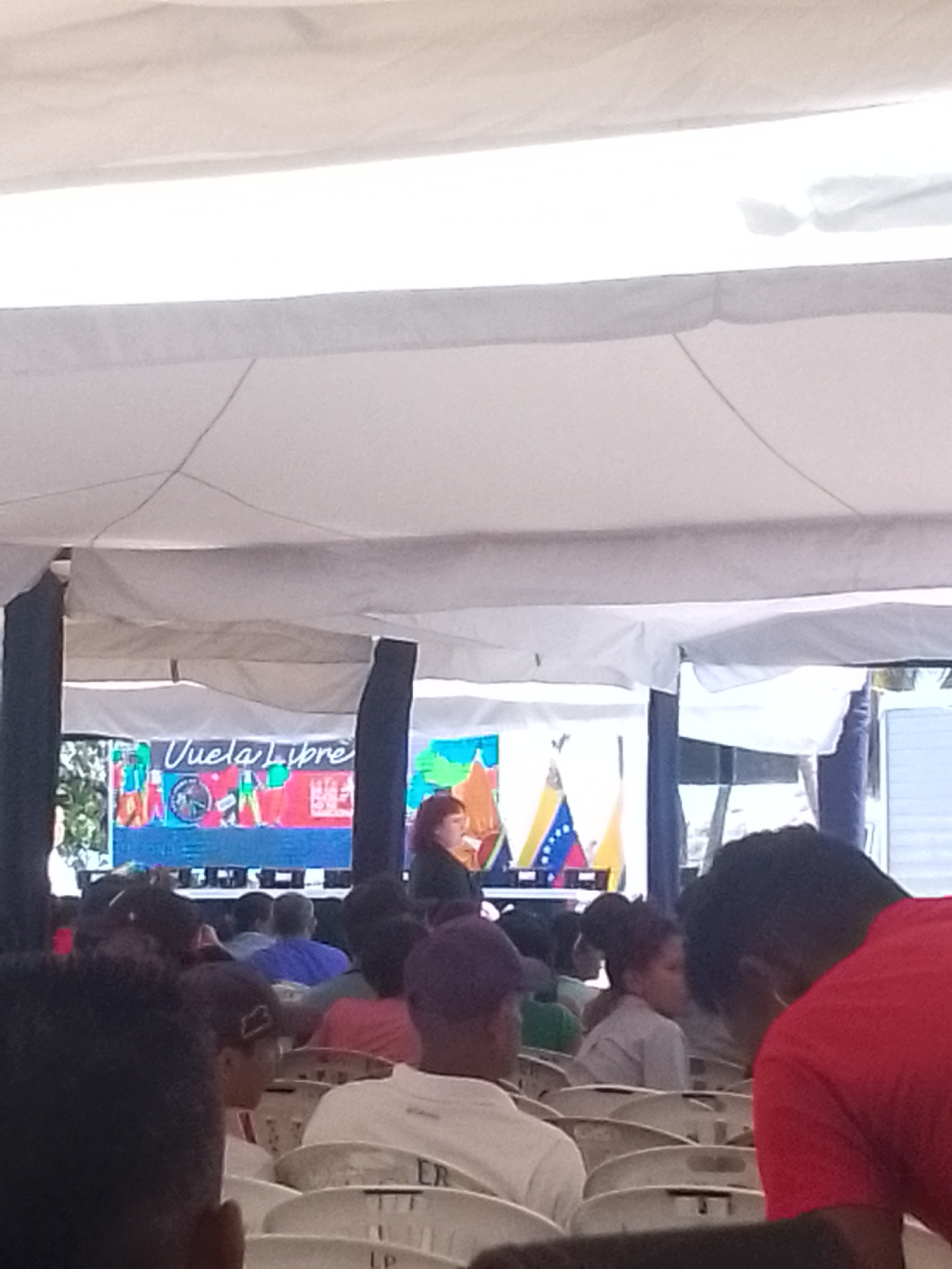 A medium indoor shot depicts a gathering of people seated in rows of white plastic chairs under a white canopy. A woman with reddish hair stands on a stage, in front of a backdrop that includes the words "Vuela Libre" in blue script and an image of the Venezuelan flag. The location is Juan Griego, Venezuela.