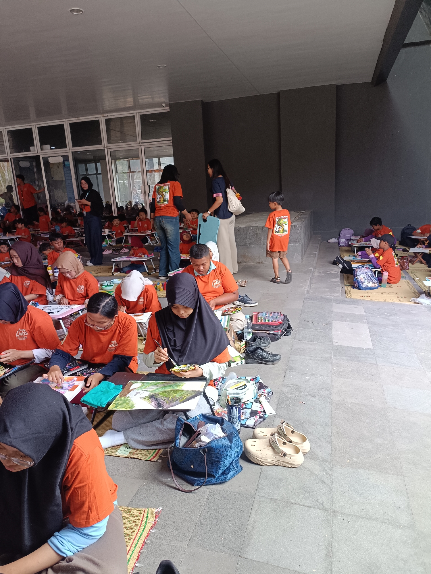 In Hargobinangun, Indonesia, a group of children and adults are gathered outdoors for an art event, possibly a drawing or painting competition. Many participants, predominantly children, are seated on the ground, arranged in rows on mats, engaged in creating art. They are mostly wearing matching orange t-shirts with a logo that appears to be "Lukis". Several adults, some in similar orange attire and others in casual clothing, are supervising or participating.
The scene takes place on a tiled outdoor floor, adjacent to a building with large glass windows and a covered overhang. Art supplies such as paint palettes, brushes, coloring pencils, and sketchpads are visible among the participants. Bags and backpacks are scattered around the area. The overall atmosphere suggests a community event focused on art and creativity.