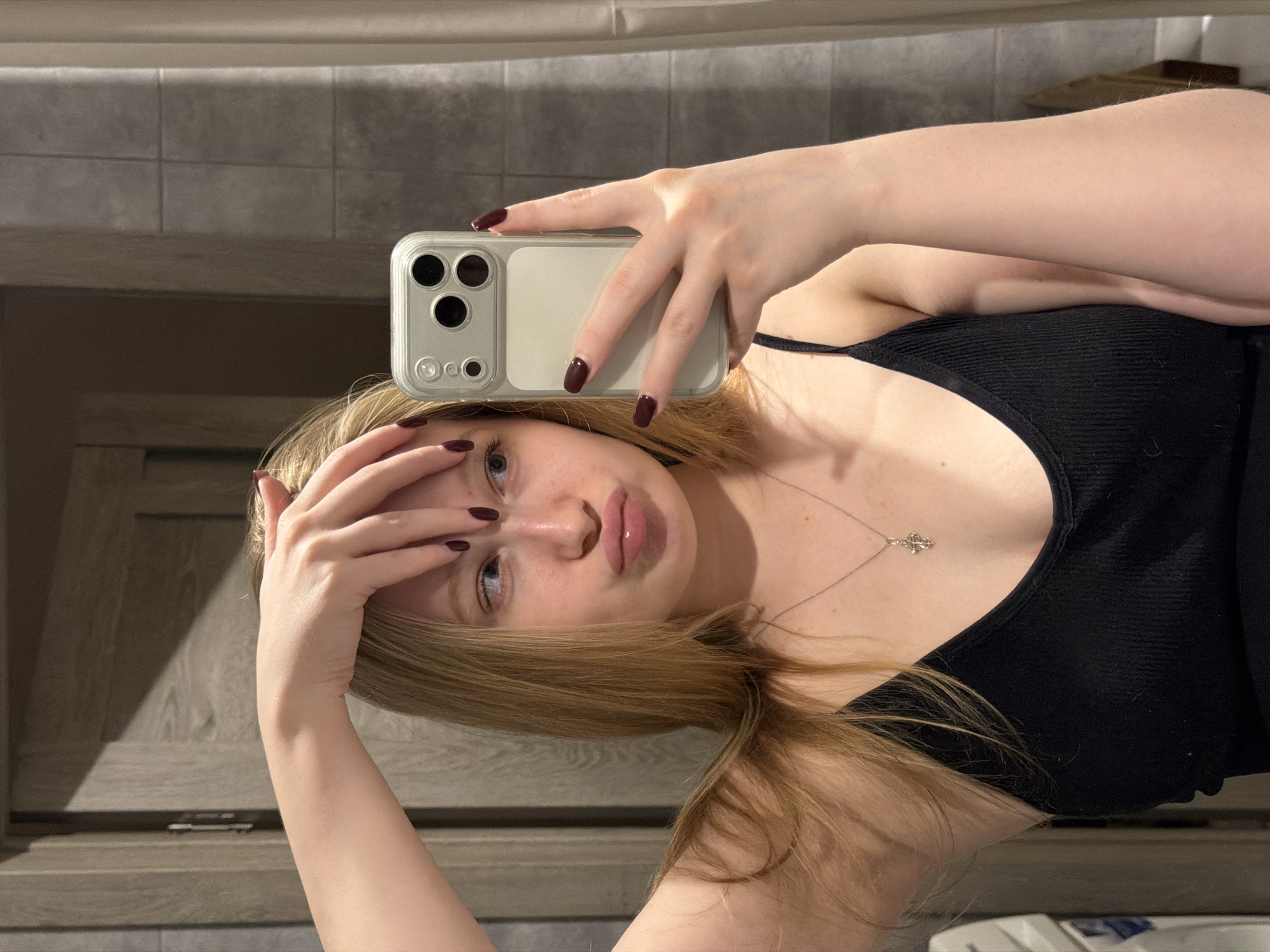 A fair-skinned woman with long blonde hair is taking a selfie indoors. She is wearing a black tank top and a silver necklace with a pendant. Her fingernails are painted dark red. She holds a white smartphone in her right hand, partially obscuring her face. Her left hand is touching her forehead. The background consists of a wall with neutral-colored tiles above and a wooden cabinet below. The image was not identified to be taken in Засечное, Russia.