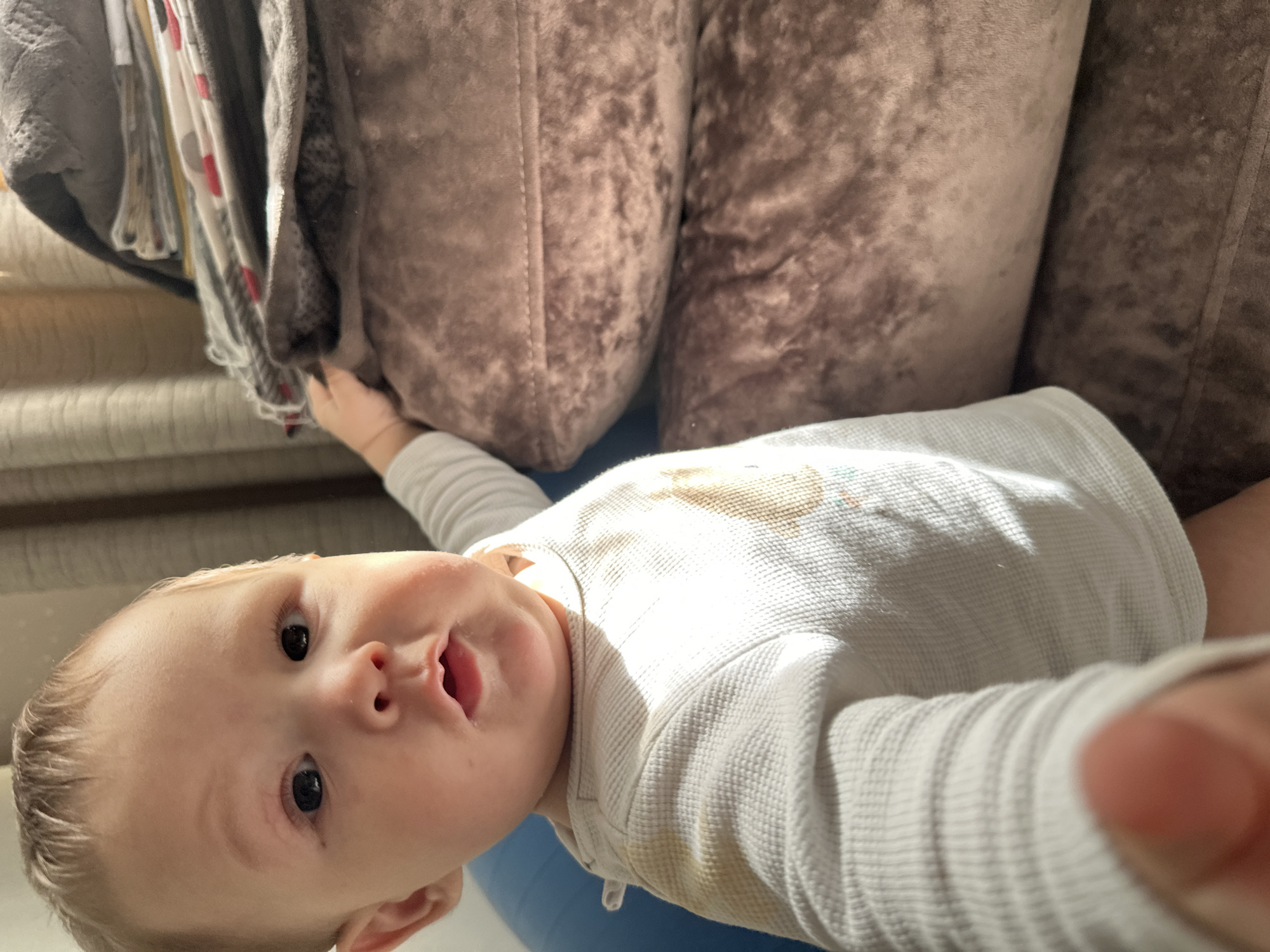 An overhead photograph captures a baby lying on its back, looking directly up at the camera. The infant has fair skin, light hair, and wide dark eyes, with an open mouth suggesting a coo or vocalization. They are dressed in a white, long-sleeved, textured thermal or waffle-knit shirt, which has some light brown or yellowish stains on the chest and right shoulder area.
One of the baby's hands is visible on the left side of the frame, reaching towards a collection of clothes hanging or draped over furniture. These clothes include a grey knitted sweater or cardigan, and a lighter fabric garment with red patterns or polka dots. To the right of the baby, and extending into the background, are two large, plush cushions from a dark brown, textured sofa or armchair. A blue exercise ball is partially visible beneath the baby, suggesting they are lying on it.
Bright, possibly natural, light illuminates the scene from the left, casting distinct shadows on the baby's face and shirt, indicating it is daytime. A blurred hand, likely belonging to the person holding the camera, is visible in the bottom right corner of the image. The baby's expression appears curious and engaged. No text is visible in the image.