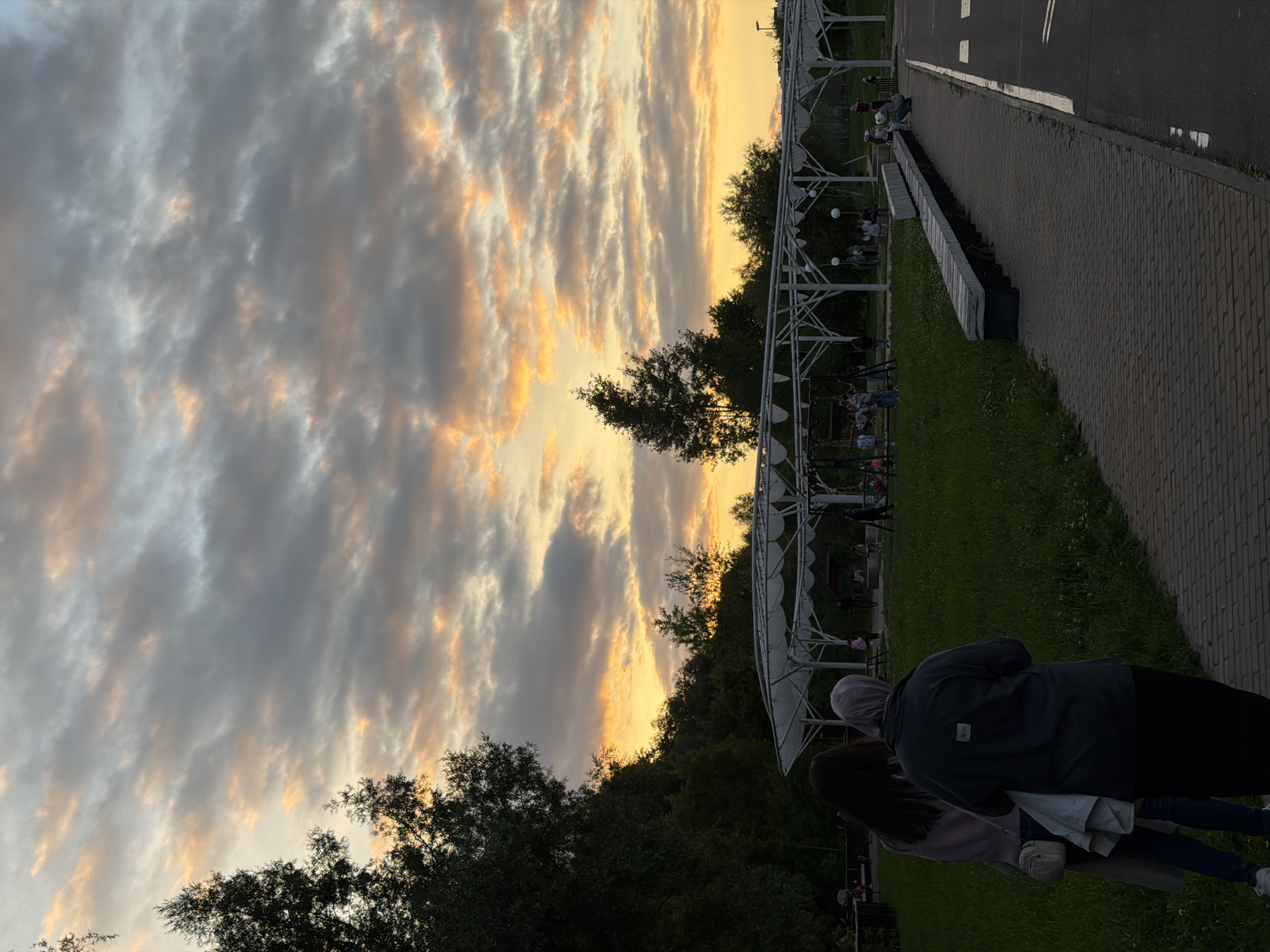 The image depicts a park scene in Kazan, Russia, during what appears to be sunset. The sky is filled with dramatic clouds, tinged with orange and yellow hues from the setting sun, contrasting with darker gray clouds. In the foreground, two women are standing with their backs to the camera, facing a grassy area. One woman has long dark hair and is wearing a light-colored coat and jeans, with a small white bag slung across her shoulder. The other woman is wearing a gray hijab and a dark long-sleeved shirt, paired with a dark skirt.
Behind them, a paved walkway runs along the edge of the grassy area. A long, dark-colored bench runs alongside the walkway. In the middle ground, there is a white, open-air structure with a curved roof supported by white pillars, resembling a gazebo or pavilion. People can be seen in and around this structure, some appearing to be children playing and adults observing. The background is populated by trees, with a dense cluster of dark green trees on the left side of the frame and scattered trees behind the pavilion. The overall atmosphere suggests a public park at dusk, with people enjoying the evening.