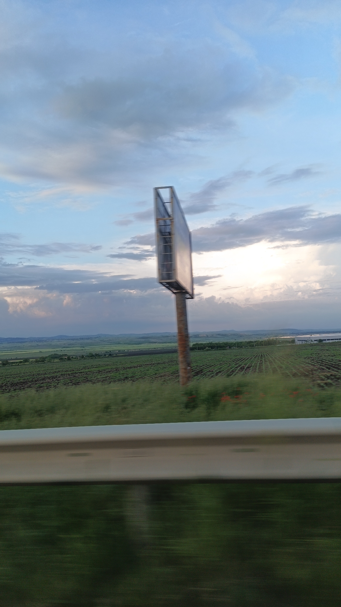 The image shows a landscape viewed from a moving vehicle, likely a car, based on the blurred foreground of a guardrail. In the distance is a rural scene. Rows of cultivated fields stretch across the landscape towards a horizon with rolling hills. A billboard stands prominently in the mid-ground, its blank surface reflecting the sky. The sky itself is filled with scattered clouds and areas of bright light. The scene appears to be on the outskirts of a city due to the cultivated fields and a warehouse in the background. Without further identifying information, it is impossible to determine the exact location in the city of Burgas, Bulgaria.