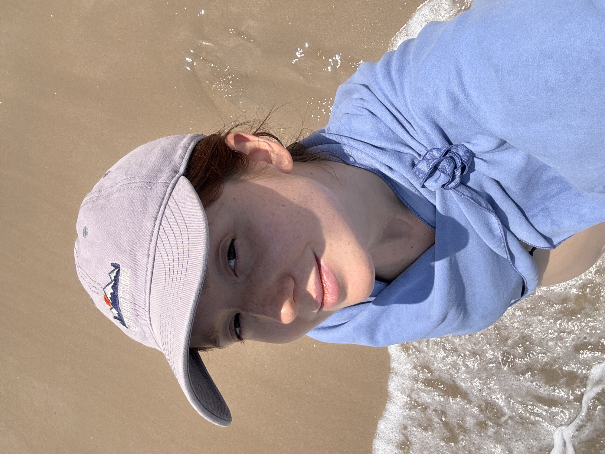 A young woman lies on a sandy beach, with the edge of the water lapping against her blue-clad shoulder. She wears a baseball cap with a mountain logo on the front. The location is likely near a beach in Choeng Thale, Thailand, based on the sand and water.