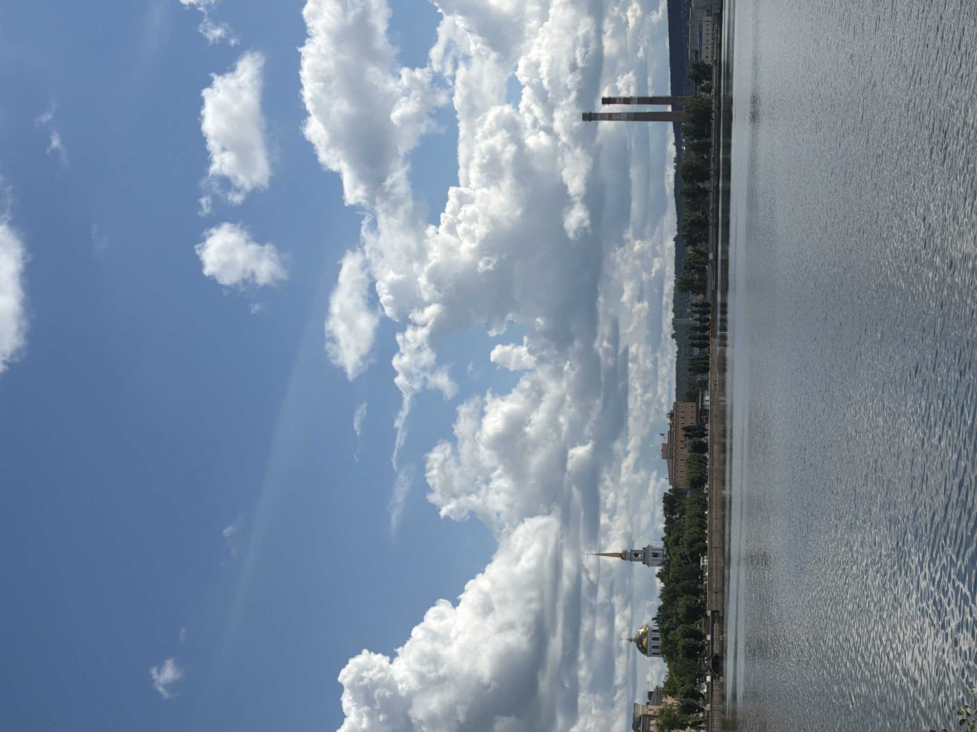 The image presents a waterfront view in Votkinsk, Russia, under a sky filled with cumulus clouds. The river (likely the Votka River) occupies the right side of the frame, displaying a ripple effect on its surface.
Along the shoreline are buildings and trees. A church with a gold dome and spire is visible, suggesting an Orthodox Christian religious building. Further along the shore, industrial structures including two tall smokestacks stand.
The sky is blue with patches of clear sky, but is mostly covered with clouds.