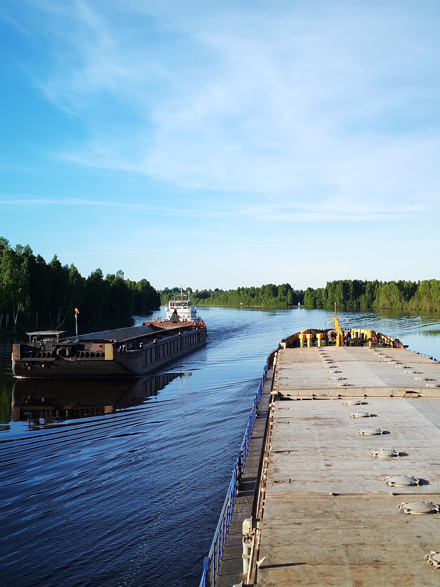 A tranquil, wide-angle photograph captures a peaceful waterway scene near Annensky Most, Russia. The perspective is from the deck of a large, flat-topped cargo barge, partially visible along the right side of the frame, featuring a blue railing and numerous circular hatches on its concrete-like surface.
In the mid-ground, a second, complete cargo vessel consisting of a dark-hulled barge being pushed by a white and red-trimmed tugboat with a prominent cabin, navigates away from the viewer. This vessel creates a distinct V-shaped wake that ripples across the calm, dark blue water. Both banks of the river are densely lined with lush, vibrant green forests, primarily composed of tall, slender trees, which are partially reflected in the water's surface.
The sky above is a bright, clear blue with sparse, wispy white clouds. The natural lighting suggests either early morning or late afternoon, as a warm, low-angle sun illuminates the trees and the visible vessel, casting subtle shadows and highlighting the textures of the water. No people are clearly discernible on either vessel or along the riverbanks. There is no legible text visible anywhere in the image. The overall atmosphere conveys serene navigation through a natural, wooded landscape.