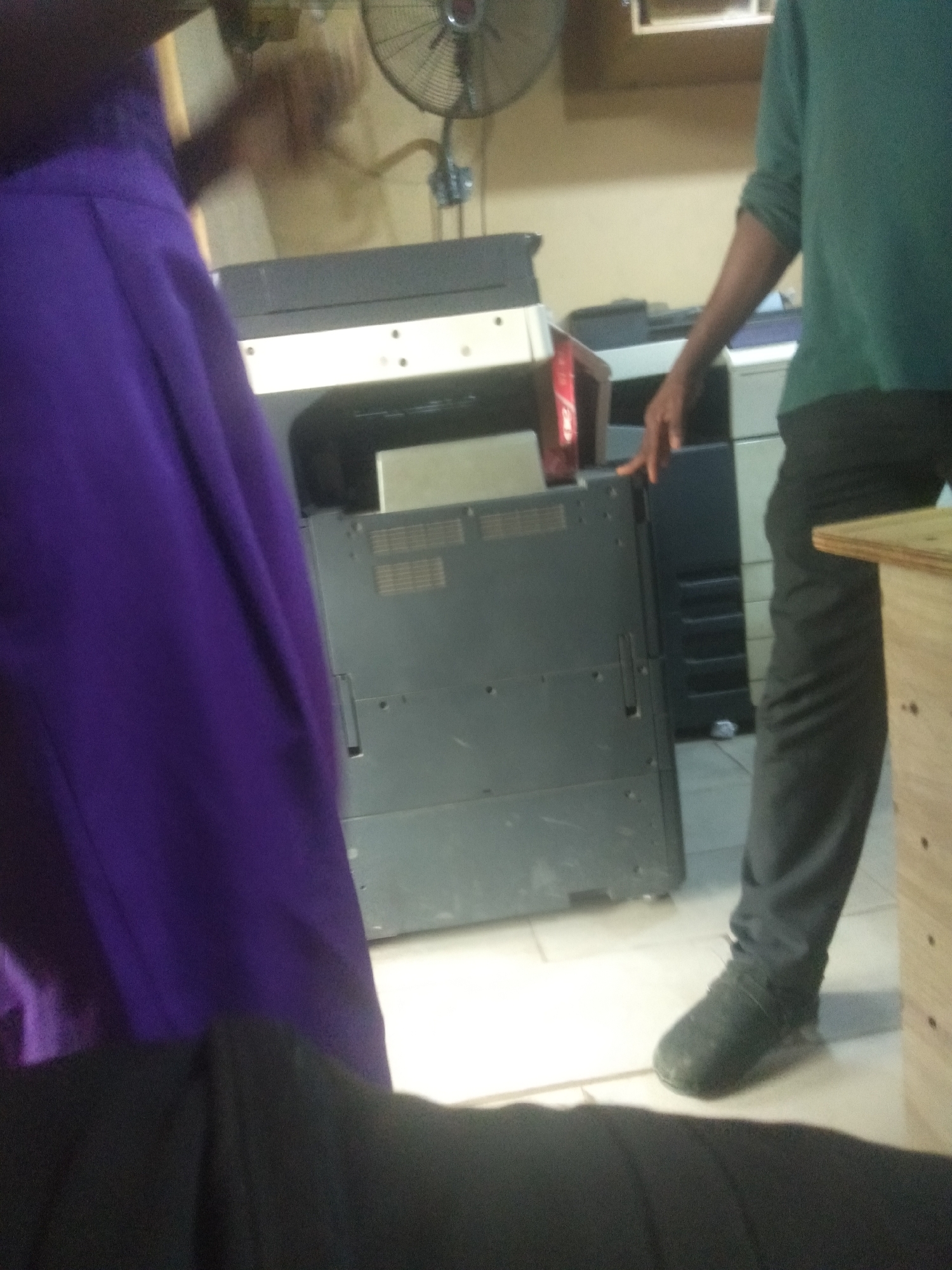 In Shangisha, Nigeria, a person dressed in a purple skirt stands beside another person wearing a green long-sleeved shirt and black pants. The second person's right hand is resting on a large, dark grey printer. The setting appears to be an office or a place with office equipment, as suggested by the presence of multiple printers and a fan in the background. The lighting is somewhat dim, and the overall scene gives the impression of an indoor environment during the day. There is no visible text in the image, and the mood appears neutral, with no strong emotional cues from the subjects. The focus of the image is on the interaction between the two individuals and the surrounding office machinery.