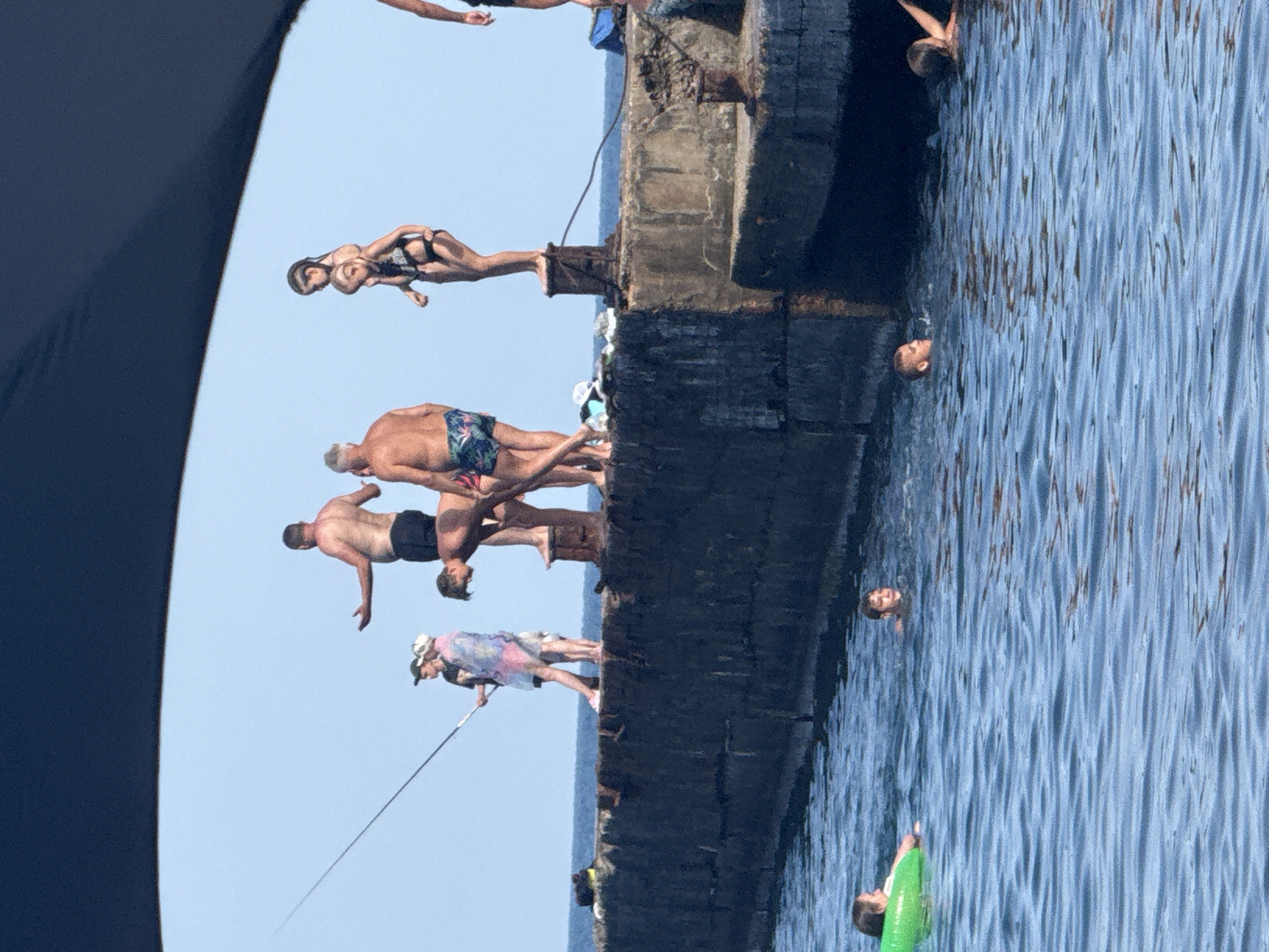 In this image, a group of people are enjoying a sunny day at a waterfront. The scene appears to be Partenit, Ukraine, with a clear blue sky overhead, suggesting pleasant weather. The main focus is on a concrete structure, possibly a pier or a wall, from which several individuals are jumping or have just jumped into the bright blue water.
Several people are captured in mid-air, demonstrating various diving or jumping poses. One person in the foreground, a woman in a black bikini, is elegantly suspended in a dive. Below her, a man in patterned swim trunks is also in the process of jumping. Further down, a man with a muscular physique is about to enter the water. Another individual, wearing a colorful garment and a hat, is positioned on the edge of the structure, holding a fishing rod, suggesting a dual purpose for the location.
In the water, several heads are visible, indicating that others are already swimming or floating. One person is holding onto a green inflatable ring. The water is calm with gentle ripples reflecting the sunlight. The concrete structure itself appears weathered, with visible textures and some signs of wear. The overall atmosphere is one of leisure and recreation. There is no visible text in the image.
