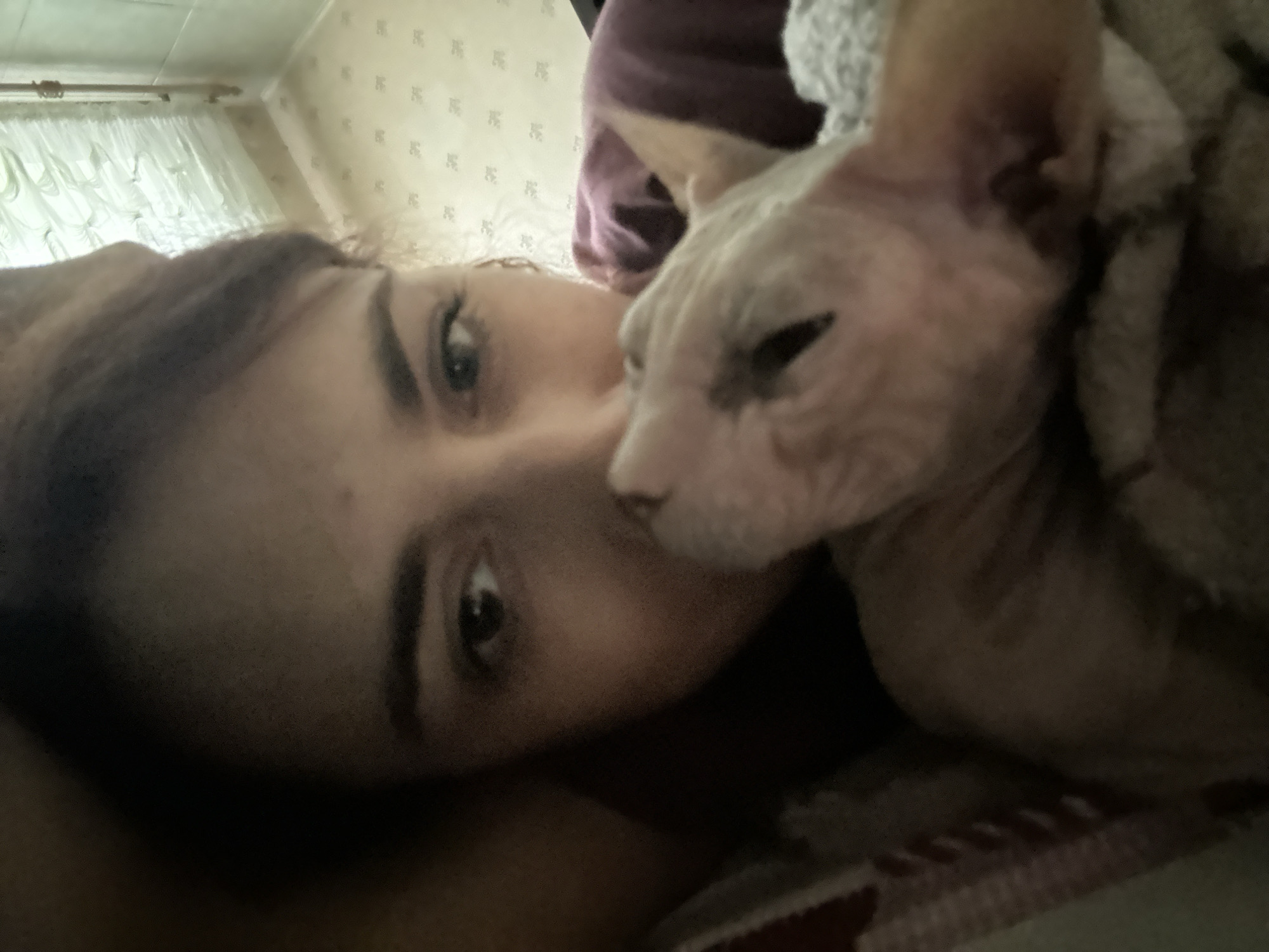 The image is a close-up selfie, taken indoors, likely in a bedroom given the presence of a bed with patterned bedding and curtains visible in the background. The lighting suggests it might be daytime, with light coming from a window.
The primary subjects are a person and a Sphynx cat. The person's face is visible, with dark hair and expressive eyes. They are positioned close to the camera, creating an intimate feel. To the right of the person, a Sphynx cat is nestled against them, with its head resting near the person's face. The cat appears to be light-colored, with wrinkled skin characteristic of the breed. The person is wearing a dark-colored top, and the cat is partially covered by a textured, light-colored blanket or piece of clothing.
The activity appears to be a moment of relaxation and affection between the person and their pet. The composition and the soft focus contribute to a sense of closeness and comfort. The background shows a wall with a subtle, repeating pattern, possibly wallpaper, and a white curtain obscuring a window.
There is no visible text in the image. The overall mood conveyed is one of warmth and connection. The location context of Togliatti, Russia, doesn't offer specific visual cues within the image itself, but implies the general setting.