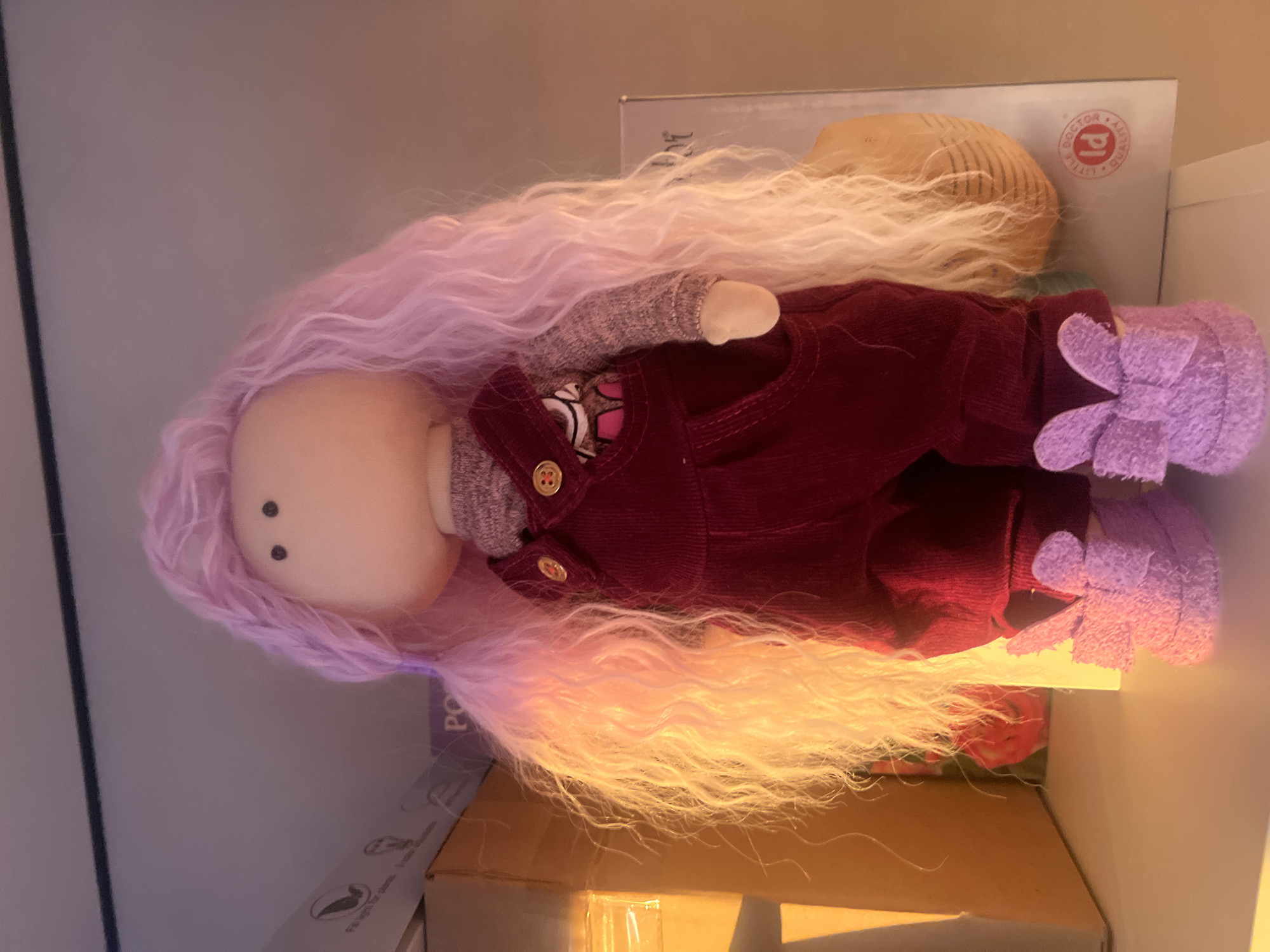 The image shows a cloth doll with a light pink head, a gray sweater, red overalls with buttons, and purple flower-shaped shoes. The doll has long, wavy hair that transitions from light pink at the top to light yellow/white at the bottom. The doll appears to be lying on a box. The presence of the doll does not provide enough information to discern the location of Votkinsk, Russia.