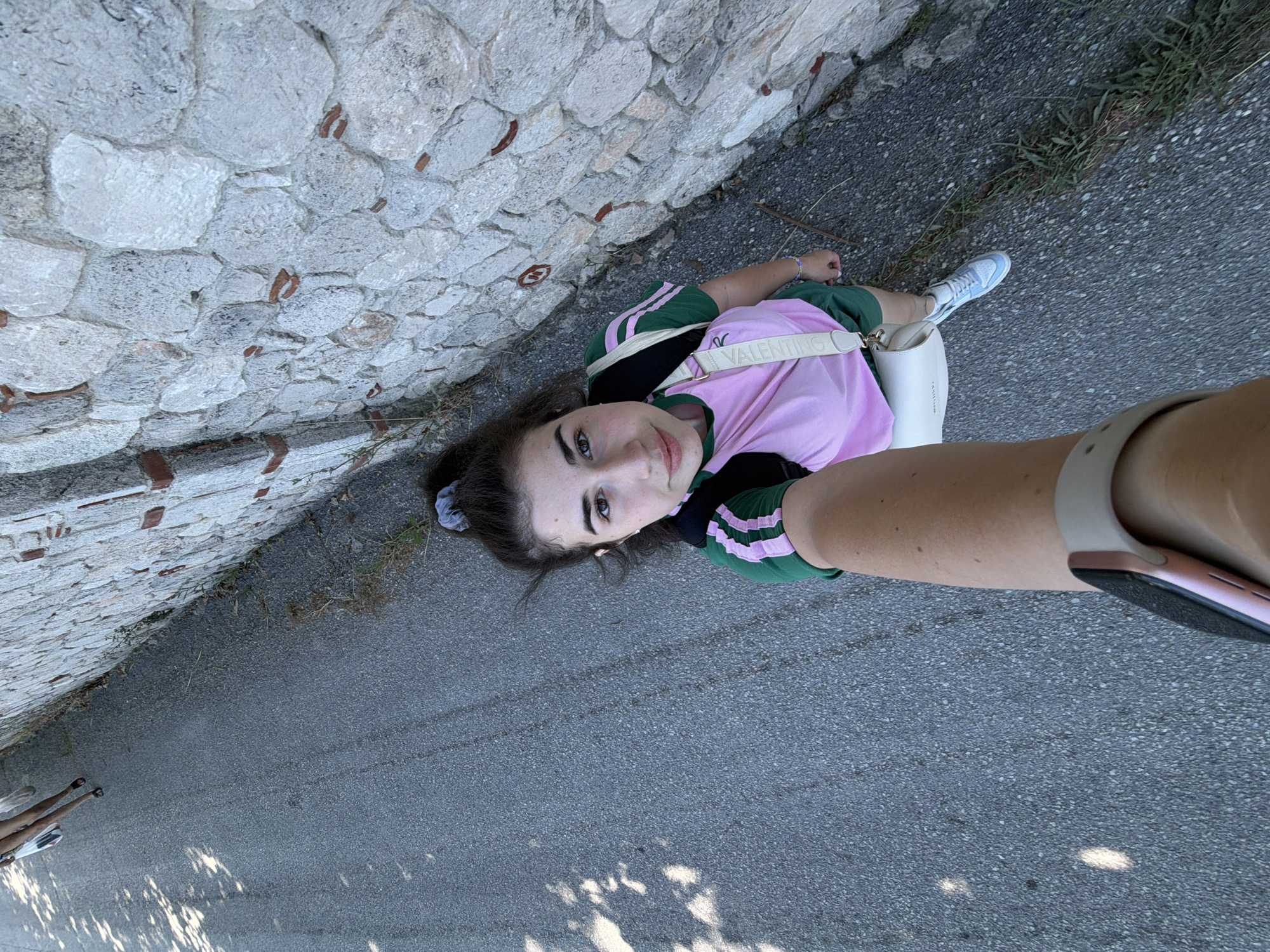 A young woman with dark hair pulled into a ponytail is taking a selfie outdoors. She is wearing a green and pink t-shirt, green shorts, and light blue sneakers. A white bag is slung over her shoulder with a strap bearing the word "VALENTINO". She is standing on an asphalt surface next to a stone wall. Sunlight casts shadows on the ground. In the background, two other figures can be seen walking away from the camera. The scene appears to be a street or pathway in a coastal area. The stone wall suggests an ancient or historical setting, possibly within the municipality of Kassandra, Greece.