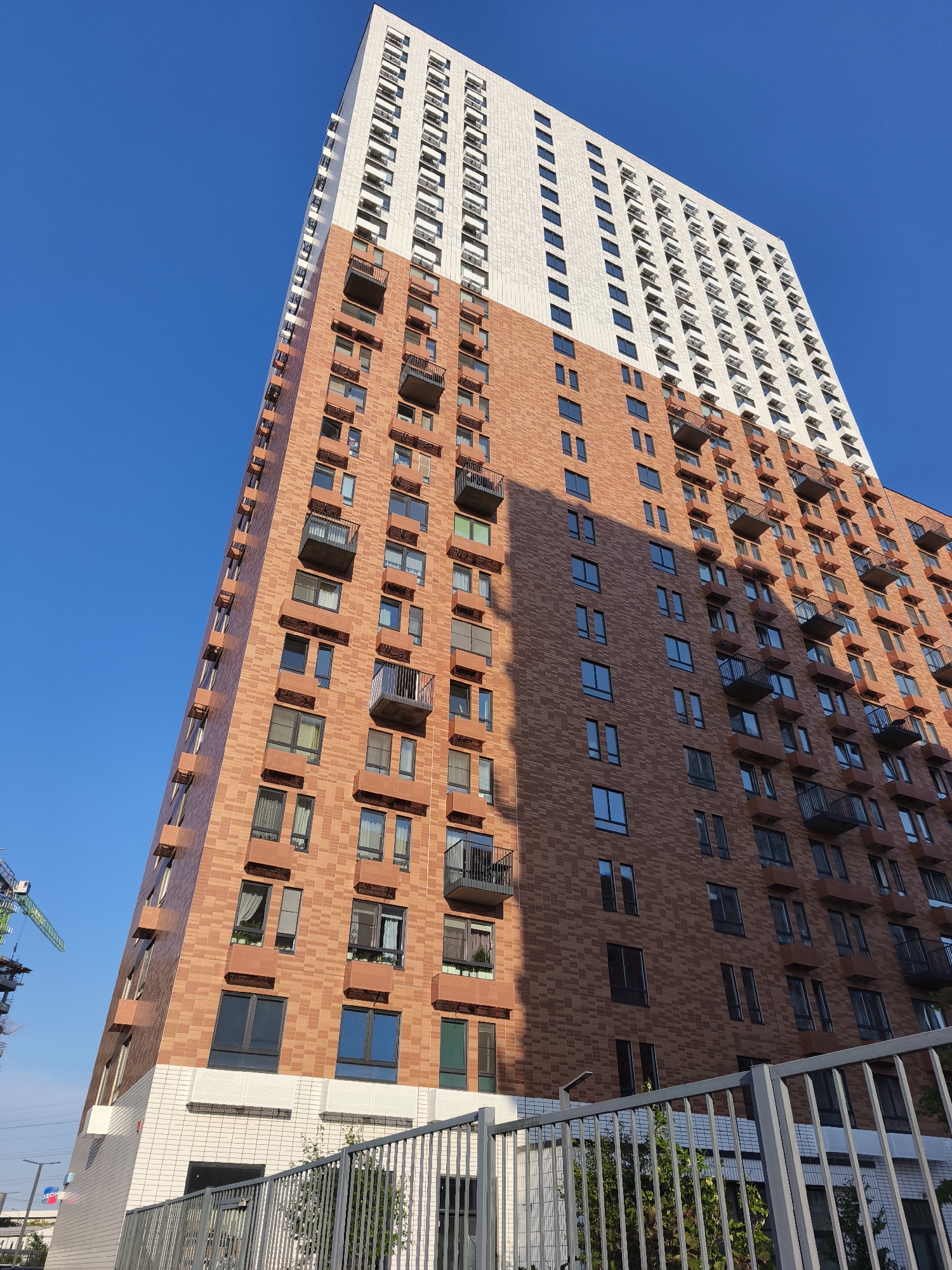 The image shows a tall residential building with a light-colored top section and a terracotta-colored lower section. The building has many windows and balconies. A construction crane is visible in the background to the left. A metal fence runs along the bottom of the image in front of the building. The sky is clear and blue. The building is located in Moscow, Russia, specifically at 19 Builders Street, Moscow.