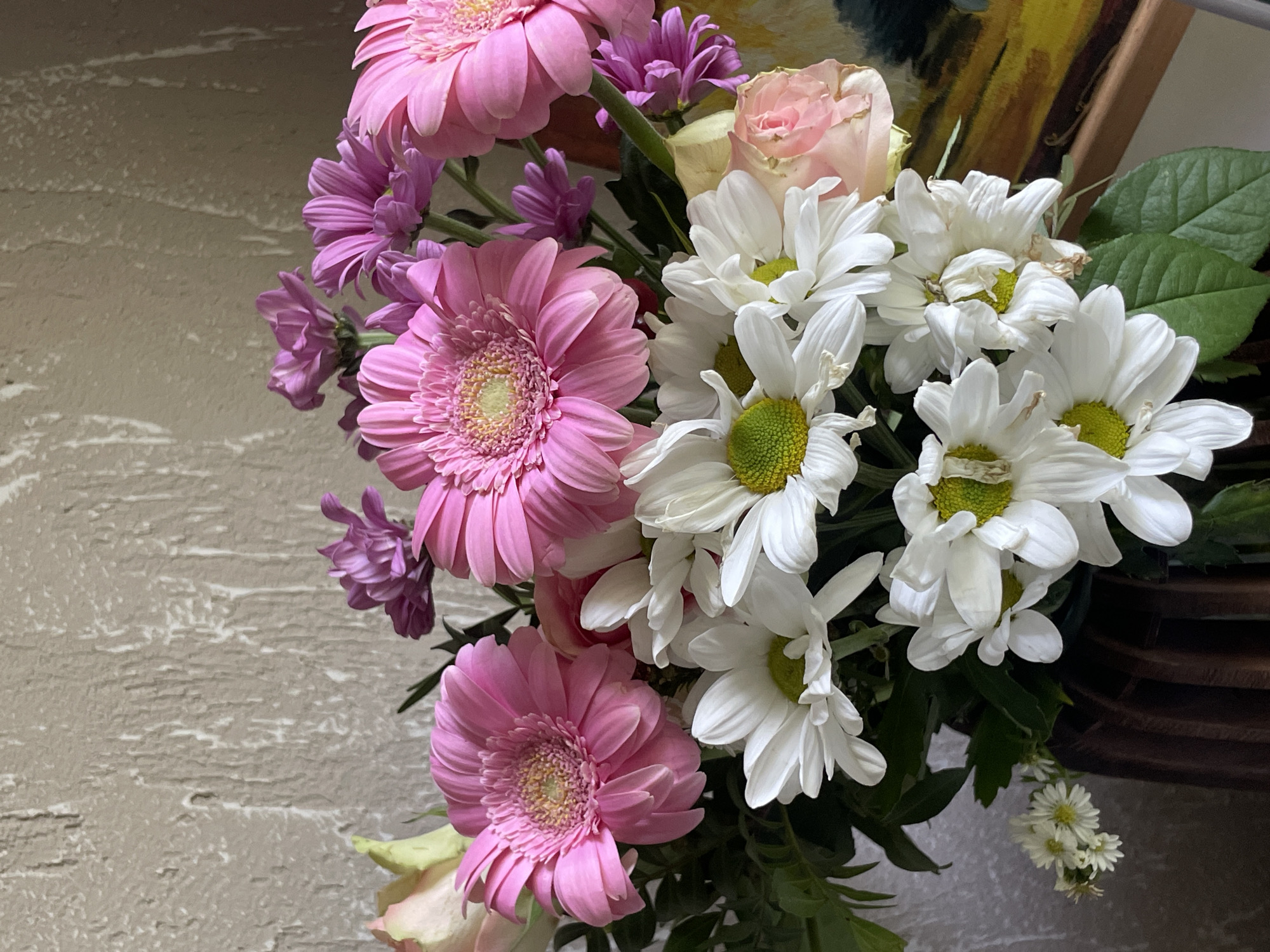 A close-up, eye-level shot displays a vibrant bouquet of flowers in a dark wooden vase. The arrangement features large, pink gerbera daisies and smaller white daisies with green centers, interspersed with delicate purple asters and a pale pink rose. Green foliage provides a lush backdrop. The flowers are positioned in the foreground, with a textured, light-colored wall behind them. To the right, a portion of a framed painting with bold yellow and dark colors is visible, leaning against the wall. The lighting suggests an indoor setting, casting soft shadows and highlighting the textures of the petals and the wall.