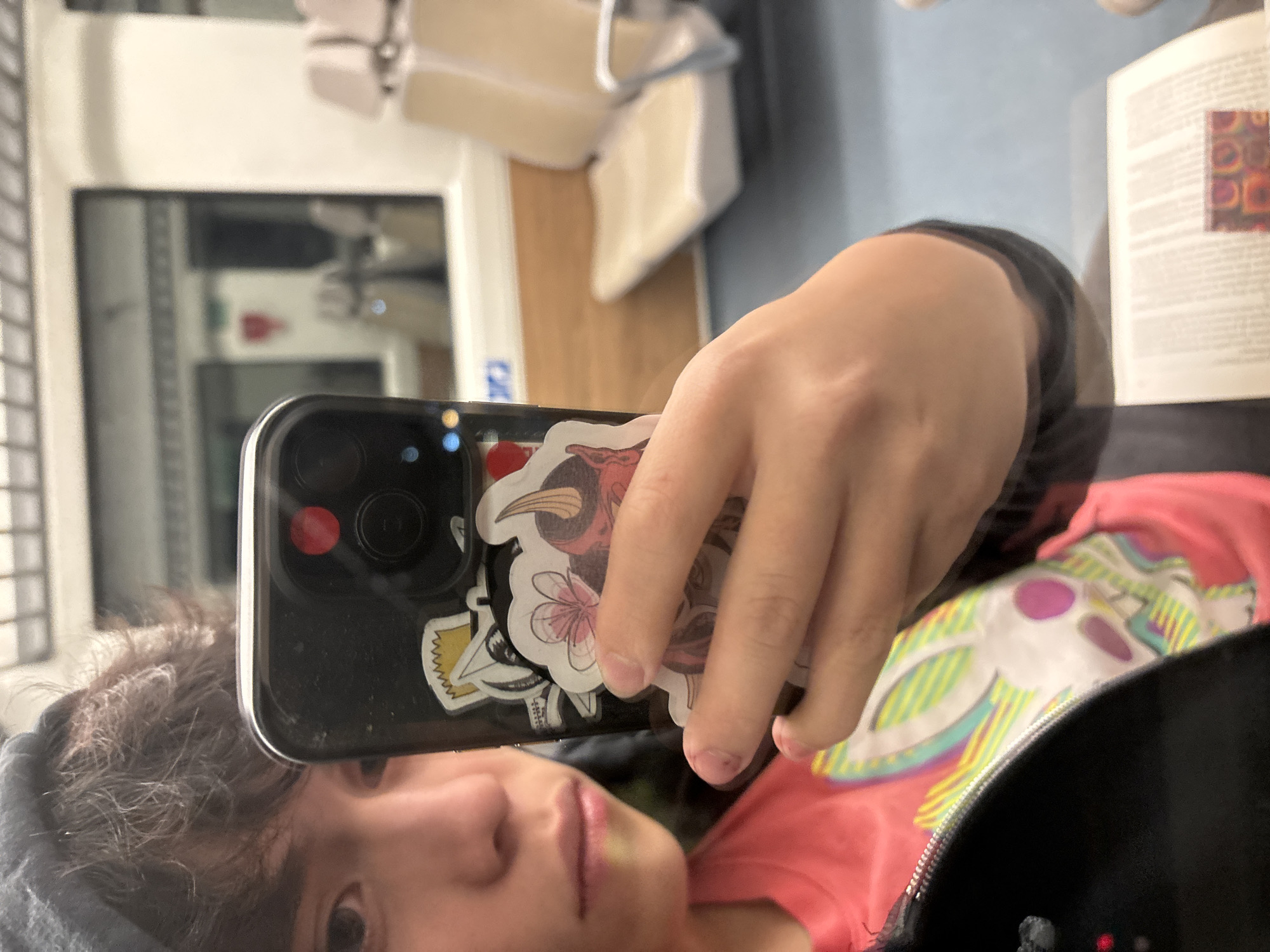 A person wearing a gray beanie, pink top and black jacket is holding a black iPhone with stickers on the back. The iPhone is angled to show the person's face in the reflection of the screen. The background shows a book, an interior space with light blue walls, an off-white chair, and a window. The location is unidentified and not necessarily in Metepec, Mexico.