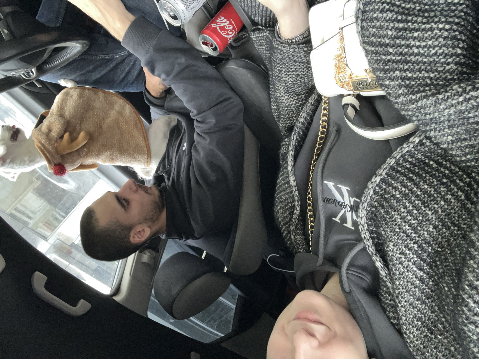 The image captures a casual scene inside a vehicle, likely a car, during the daytime.
On the left side, a man with short dark hair and a beard is seated, wearing a dark long-sleeved top. His left arm is visible, resting on his leg. Beside him, a small white dog is perched, wearing a light brown, fluffy reindeer-themed hat complete with small antlers and a red pom-pom nose. The dog appears to be looking out a window.
On the right side, a woman is partially visible, with her lips and torso in view. She is wearing a dark grey hoodie with "Calvin Klein Jeans" visible on it, layered under a black and white patterned knit cardigan or jacket. A white handbag with a gold chain strap and gold embellishments rests on her lap, displaying the text "VERSACE JEANS COUTURE."
Between the two people, a red "Coca-Cola" can and another silver can are placed, likely in a cup holder or on a console. The car interior features dark seats and an overhead handle. Bright natural light illuminates the scene through the car windows, suggesting it is daytime. The blurred background seen through the windows indicates an outdoor setting, consistent with Varna, Bulgaria. The individuals appear to be passengers, or one is a driver, during a journey or a stop.