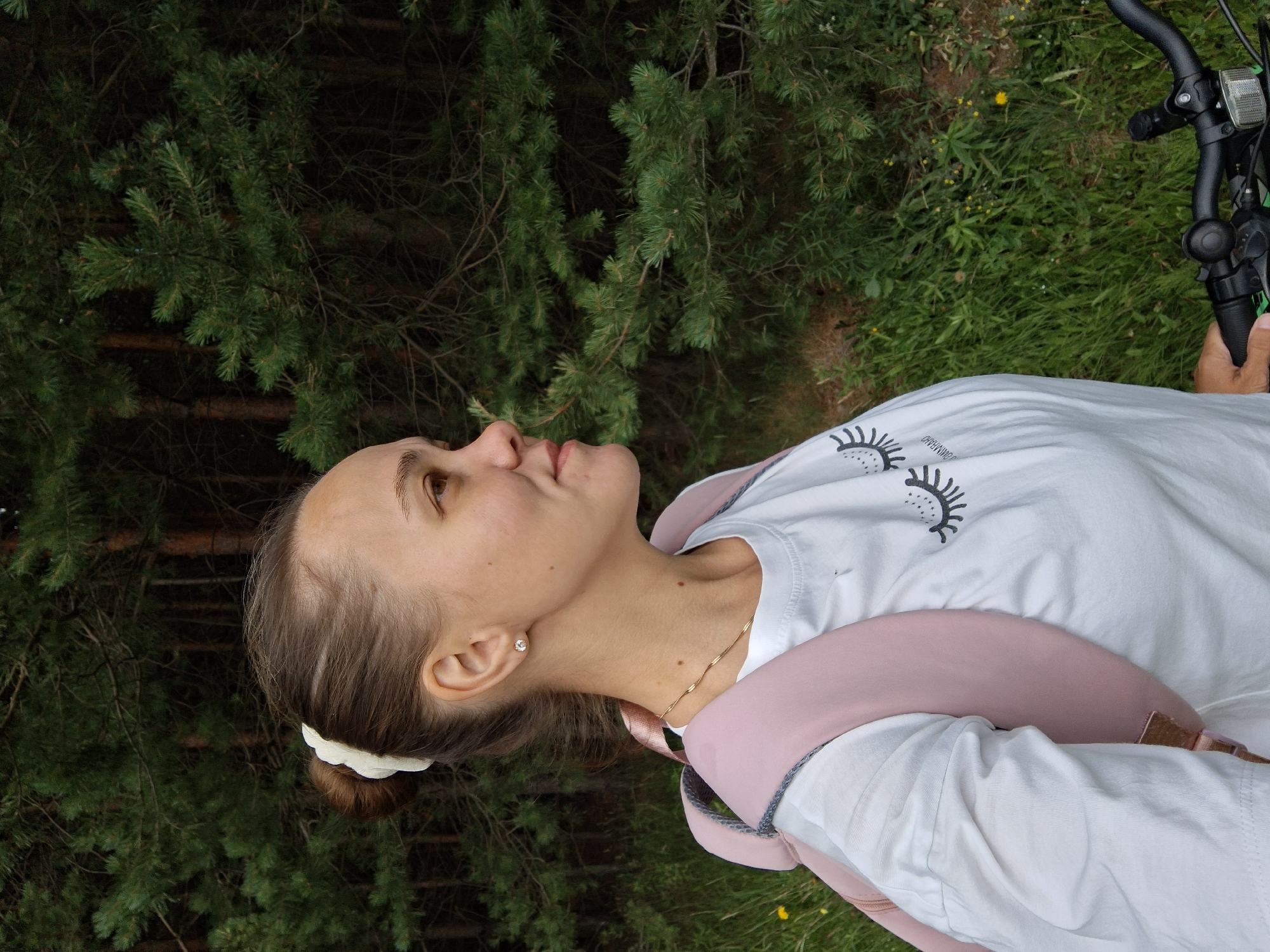 A young woman, viewed from the chest up, is in profile, looking upwards and to her left. She has her hair pulled back into a bun with a white scrunchie and is wearing a white t-shirt with a graphic of eyelashes. A pink backpack is slung over her shoulders. Her skin is fair with a few freckles visible on her nose and cheeks. She is wearing a small diamond stud earring and a delicate gold necklace. The background is out of focus, showing green pine trees and grass, suggesting an outdoor setting. The lighting appears to be natural daylight. The image is oriented vertically, with the woman's head at the top. The scene is peaceful, with the woman appearing contemplative or perhaps enjoying the view. The image appears to have been taken in Chaikovsky, Russia.