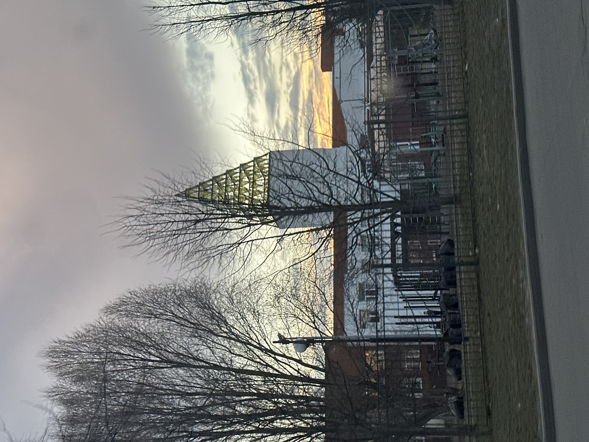 The image shows a view of the Shukhov Radio Tower in Moscow, Russia. The tower, a lattice steel structure, is partially obscured by bare trees. The background includes a building and a cloudy sky with a hint of sunset. A grassy area and a road border the right side of the frame.