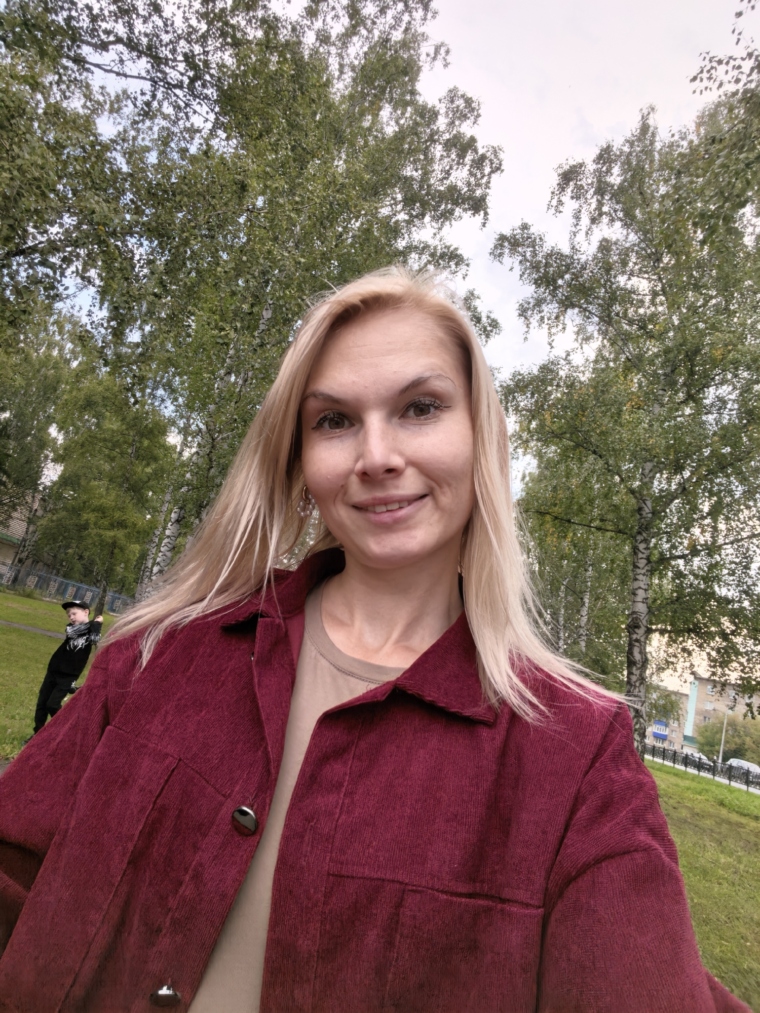 A blonde woman in a maroon corduroy jacket smiles at the camera, taken from a low angle. Behind her, a child in dark clothing with a scarf and hat is visible in the background, walking on a grassy area. Trees with green foliage and birch trees are prominent in the background, with a glimpse of buildings and a street in the distance, suggesting an urban park or public space in Sterlitamak, Russia. The sky is overcast.