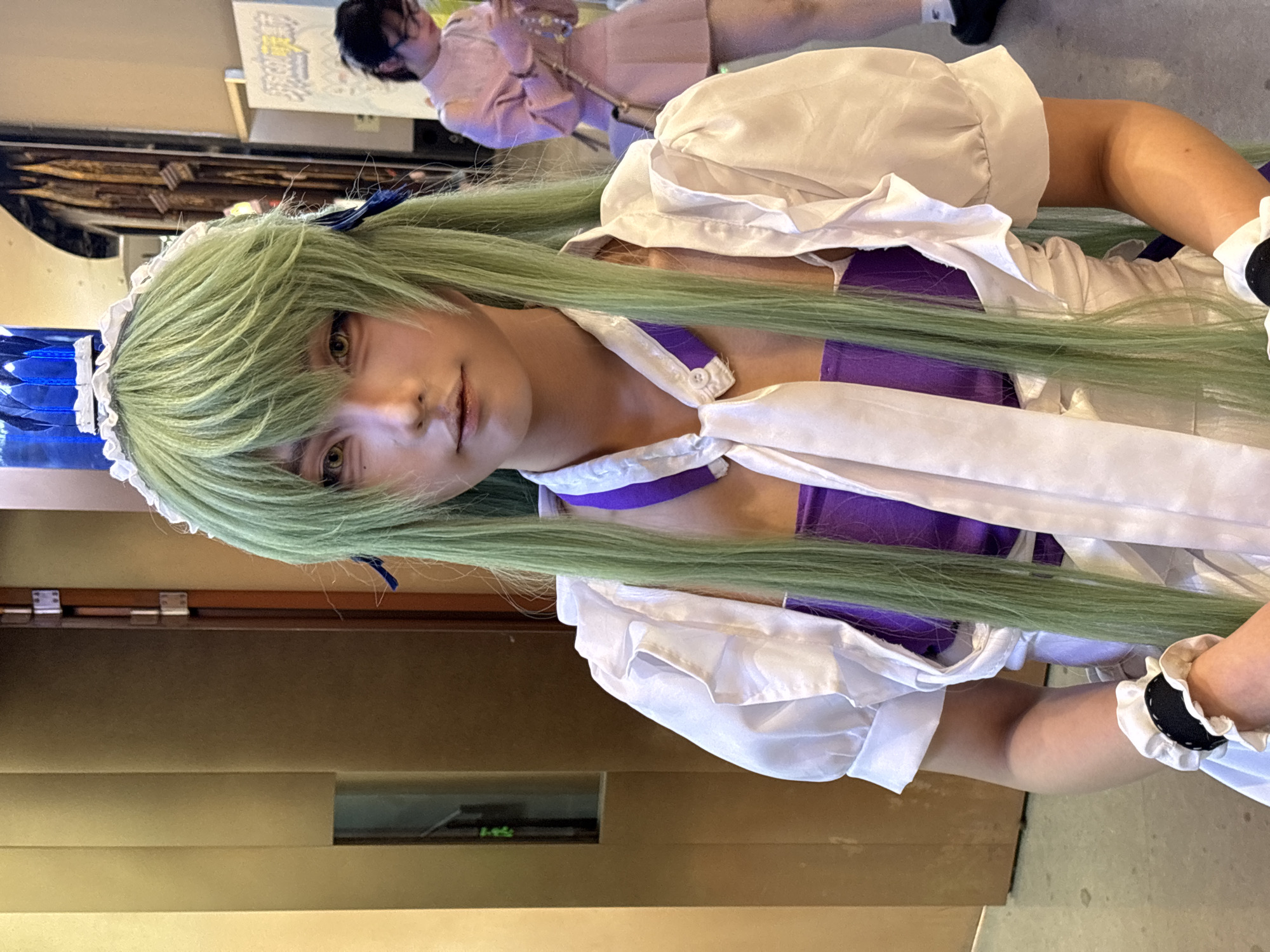 A person with green hair in a white and purple costume is centered. The person is wearing a blue and white headdress. Their costume has a white collar and a purple underlayer. A white ribbon is tied at the collar. The person has fair skin and light brown eyes.
In the background, another person is playing a flute. A sign is visible in the upper left of the frame. A brown and gold wall is in the lower left. The location is unknown.