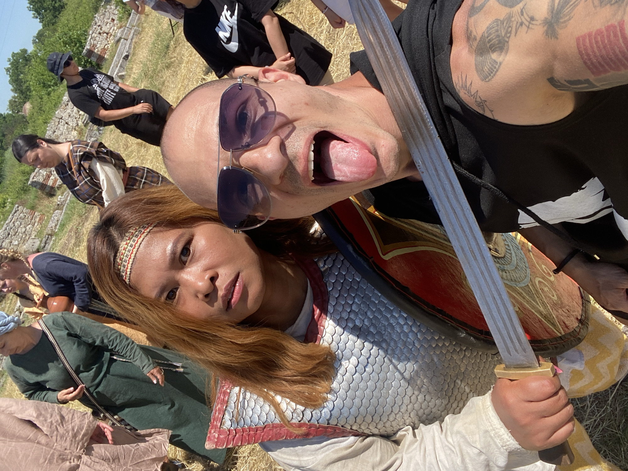 The image captures an outdoor scene at the archaeological site of Oescus in Gigen, Bulgaria, under sunny conditions. In the foreground, two individuals are depicted. On the left, a female subject, appearing to be of East Asian descent, has long, reddish-brown hair adorned with a multicolored patterned headband. She is wearing a white long-sleeved tunic beneath a silver scale armor (lorica squamata) with red trim. She holds a circular shield, predominantly red with gold and blue decorative patterns, and a brass-hilted sword in her right hand. Her expression is directed towards the camera with a neutral gaze. To her right, a male subject with a shaved head and multiple tattoos on his right arm is wearing dark aviator sunglasses with purple lenses and a black sleeveless hooded top. His mouth is open, with his tongue extended. The sword's blade extends diagonally across his upper chest.
In the mid-ground, several other individuals are present. To the left, a person in a light brown tunic is partially visible. Adjacent to them stands an older woman wearing a long, dark green dress, a light blue head covering, and a patterned cross-body strap. Behind her, a woman in a yellow garment holds a brown clay pot. Further right, a woman with dark hair pulled back is dressed in a brown and plaid patterned garment. In the background, behind the foreground male, a person in a black t-shirt, shorts, dark bucket hat, and sunglasses stands with hands in pockets. A second person in a black t-shirt with a white "NIKE" logo is partially visible on the far right, alongside a child's arm.
The ground is covered with dry grass and straw. The background features significant stone ruins, indicative of an archaeological site. A distant hill topped with green foliage and trees is visible against a clear blue sky.