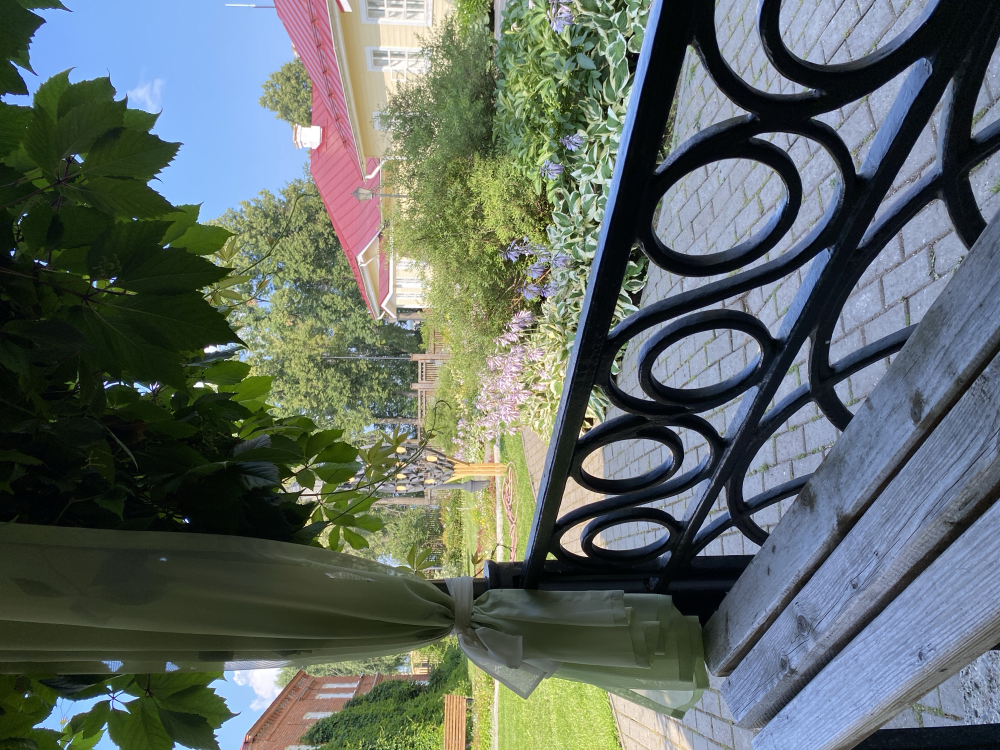 The image depicts a tranquil outdoor scene at the Museum-Estate of P.I. Tchaikovsky in Votkinsk, Russia. In the foreground, a weathered wooden bench sits before an ornate black metal fence with circular motifs. To the left, a light green sheer curtain is tied to a pole, framing a view of the estate's grounds. Beyond the fence, a paved walkway leads through a meticulously maintained garden filled with lush green foliage and clusters of purple flowers. Two yellow buildings with red roofs are visible in the middle ground, one with white-trimmed windows. A brick building covered in ivy stands to the far left, with a wooden bench visible on its lawn. The sky above is a clear blue with a few wispy clouds. The overall atmosphere is one of serene natural beauty and historical preservation.