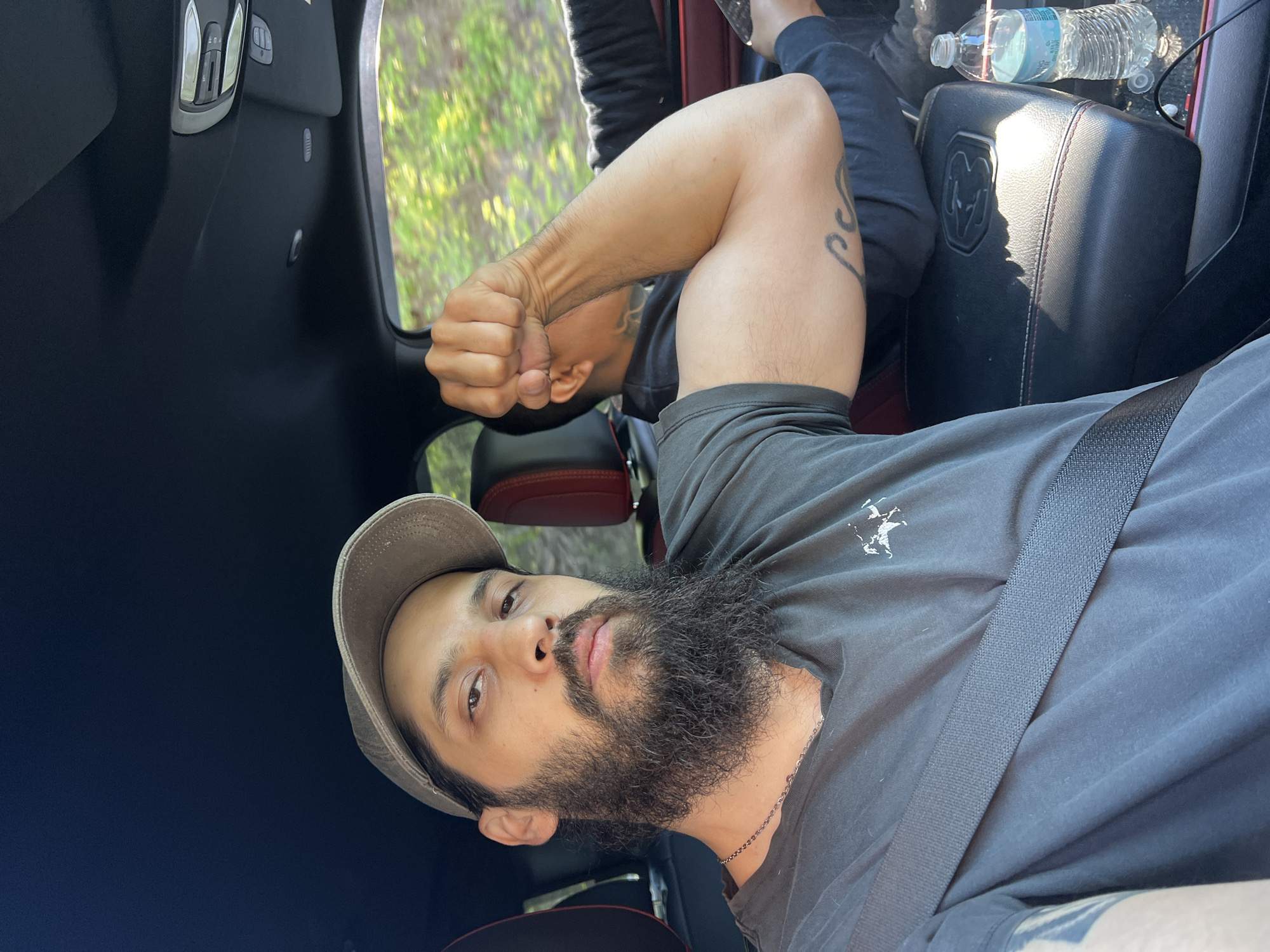 The image, rotated 90 degrees counter-clockwise, captures a man seated in the front passenger seat of a vehicle, likely in Balmville, United States of America. He looks directly towards the camera with a neutral expression, wearing a light brown baseball cap and a dark grey t-shirt featuring a small white emblem on the left chest. A black seatbelt is fastened across his torso. He has a full, dark beard and mustache.
His right arm is bent at the elbow, fist clenched, showcasing visible muscle definition and tattoos, including an elaborate design on his bicep. Another arm, presumably his left, is visible in the background, also bent and flexed.
The vehicle's interior is dark, with black upholstery and paneling. Red accents or stitching are visible on the seat or console. A prominent black headrest features an embossed "RAM" logo, indicating the vehicle is likely a Dodge Ram. A plastic water bottle with a blue label rests on the center console. Through the vehicle's windows, a blurry backdrop of green foliage suggests a natural, outdoor setting under daylight conditions.