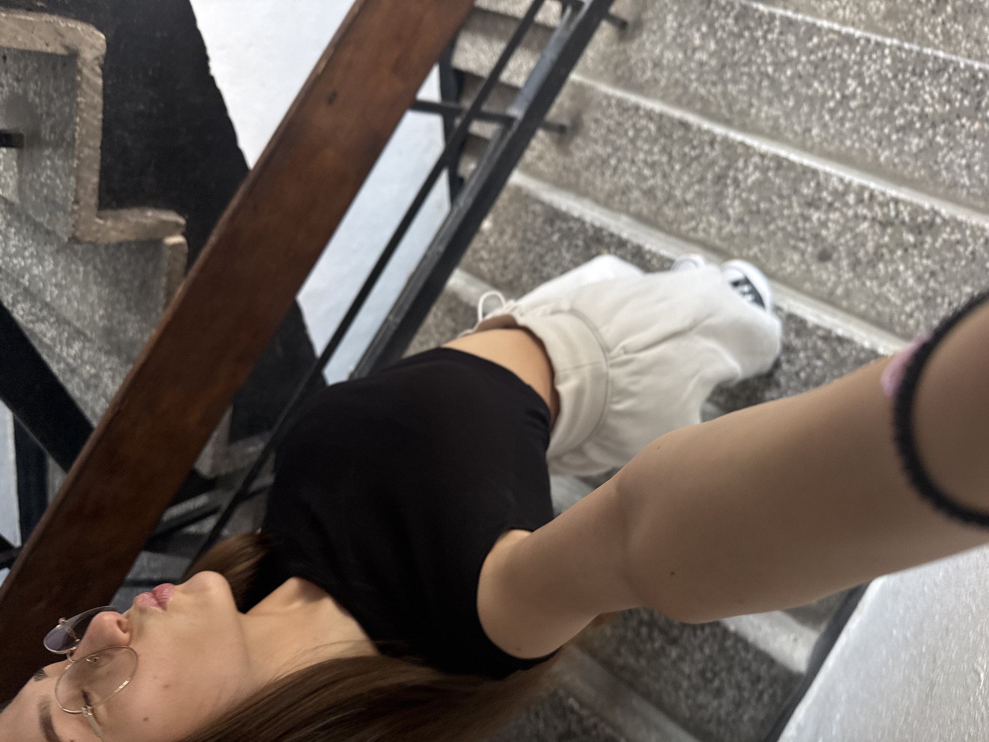 A young woman with long brown hair and glasses is posing on a concrete staircase, puckering her lips towards the camera. She is wearing a black crop top and light-colored shorts, with sneakers visible on her feet. The staircase has a wooden handrail and a black metal railing. The environment appears to be an indoor or covered outdoor staircase, possibly in an apartment building or public space.
