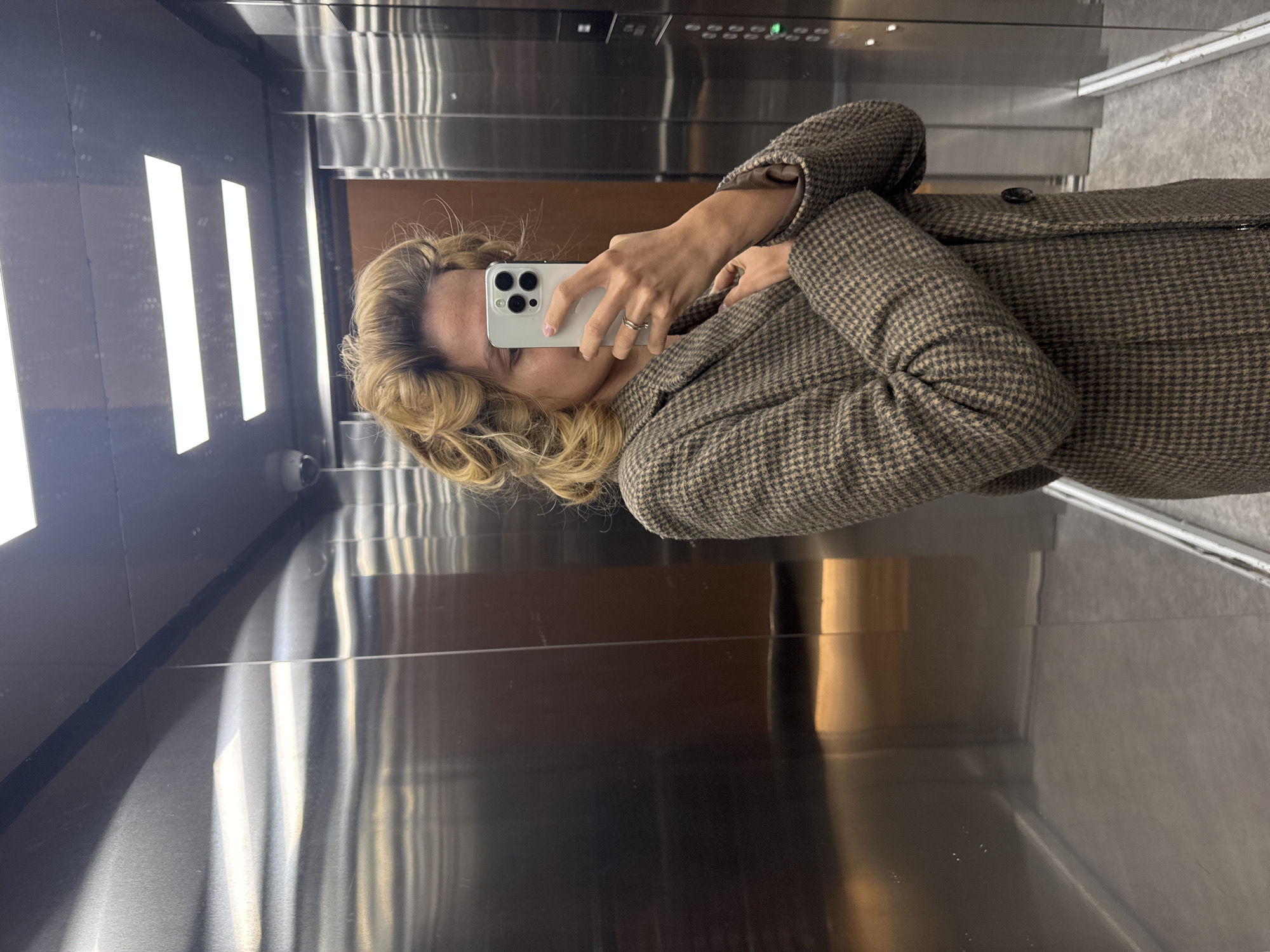 A woman is taking a mirror selfie in what appears to be an elevator. She is holding a white iPhone with three camera lenses, partially obscuring her face. She has blonde, wavy hair and is wearing a brown and tan patterned coat. The elevator interior is a mix of stainless steel and dark brown surfaces, with elongated rectangular light fixtures on one wall. Buttons and indicator lights are visible above her head on the stainless steel surface. The location is Unknown, Unknown.