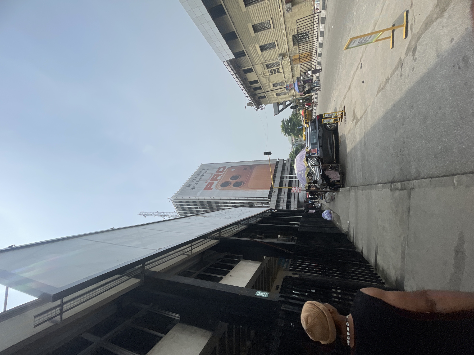 The image depicts a street scene in Lagos, Nigeria. A tall building featuring an advertisement of a mobile phone is visible in the background. A black vehicle and several pedestrian vendors with carts are along the sidewalk. One person is wearing a tan knit cap. The street has some markings, likely for vehicle guidance, and what appears to be a pedestrian traffic barrier. The buildings in the background are multistory, with windows. The sky is visible above, with a slightly overcast appearance. The foreground shows part of a dark-colored building structure.