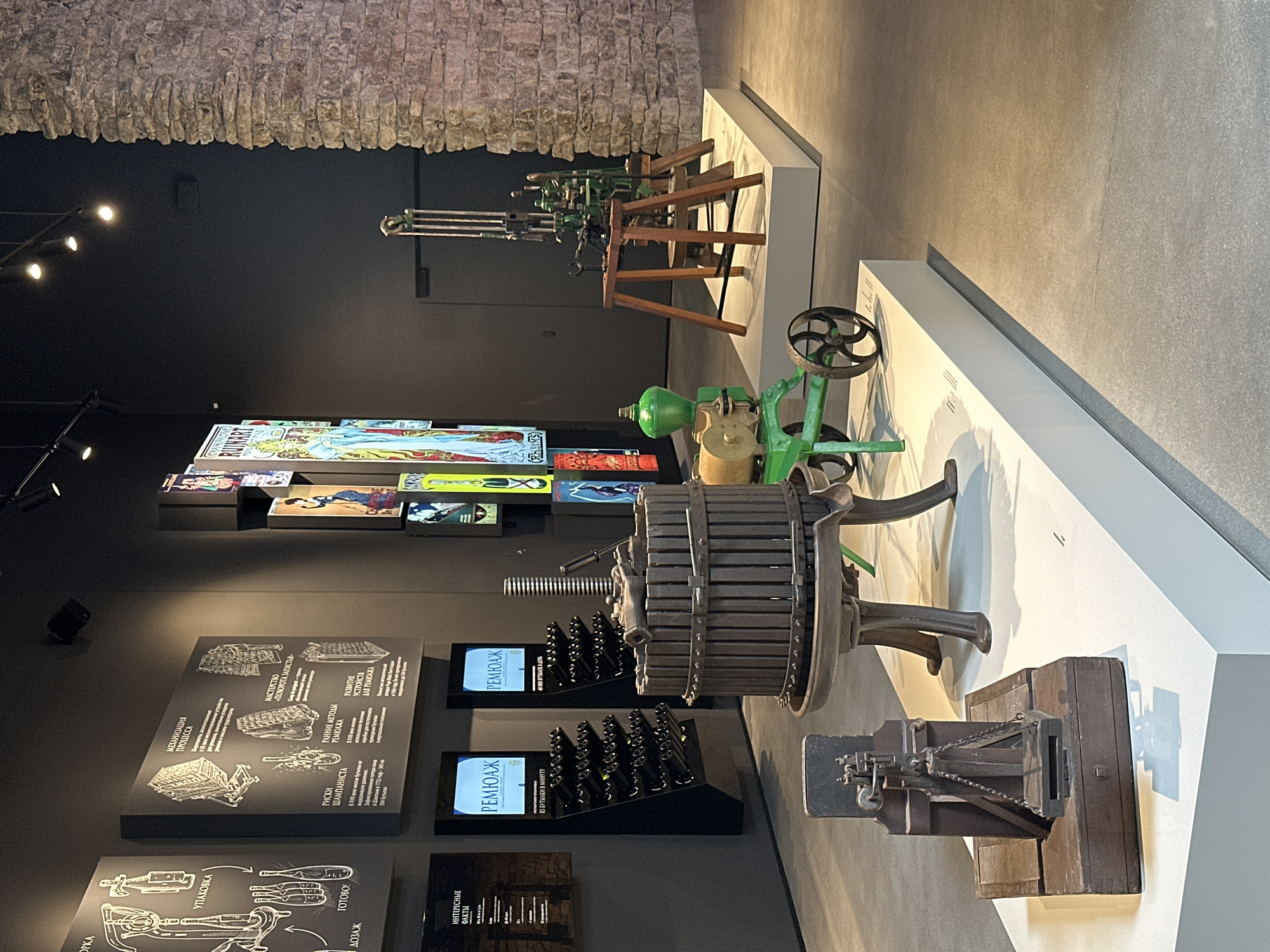 This media file depicts an indoor exhibit, likely a museum or historical display, focused on the history of wine production, specifically champagne. The scene is dimly lit, with spotlights highlighting various artifacts.
The main subjects are several historical machines and displays. In the foreground, a large, wooden wine press is prominently featured. To its right, a smaller, intricate metal machine with chains sits on a wooden base. In the background, a green and metallic machine with a flywheel and a hopper is visible. Further back, a series of wooden frames hold rows of metal components, possibly part of an assembly line or a different type of manufacturing equipment.
The exhibit includes educational panels with illustrations and text. One panel, in the lower left, illustrates the process of "УПАКОВКА" (packaging) and "ДОЗАЖ" (dosing). Another panel describes "МЕХАНИЗАЦИЯ ПРОЦЕССА" (mechanization of the process) and details "МАСТЕРСТВО «ПОВОРОТА ЗАПАСТЬЯ»" (mastery of "turning the neck"). It also mentions "РИСКИ ШАМПАНИСТА" (risks of the champagne maker) and "РАННИЕ МЕТОДЫ РЕМЮАЖА" (early methods of riddling), along with "РАЗВИТИЕ УСТРОЙСТВ ДЛЯ РЕМОНЖА" (development of riddling devices). Text on the display stands reads "РЕМЮАЖ" and indicates "83 БУТЫЛКИ В МИНУТУ" (83 bottles per minute) and "40 000 БУТЫЛОК В ДЕНЬ" (40,000 bottles per day). Another panel has the title "ИНТЕРЕСНЫЕ ФАКТЫ" (interesting facts). Colorful posters or artwork, possibly advertisements or historical illustrations, are displayed on the wall in the mid-ground.
There are no people visible in the image. The setting appears to be a modern exhibition space with dark walls and white display platforms, contrasted with the rough, textured stone ceiling or upper wall, suggesting an older architectural element incorporated into the design. The overall impression is one of a well-curated historical display.