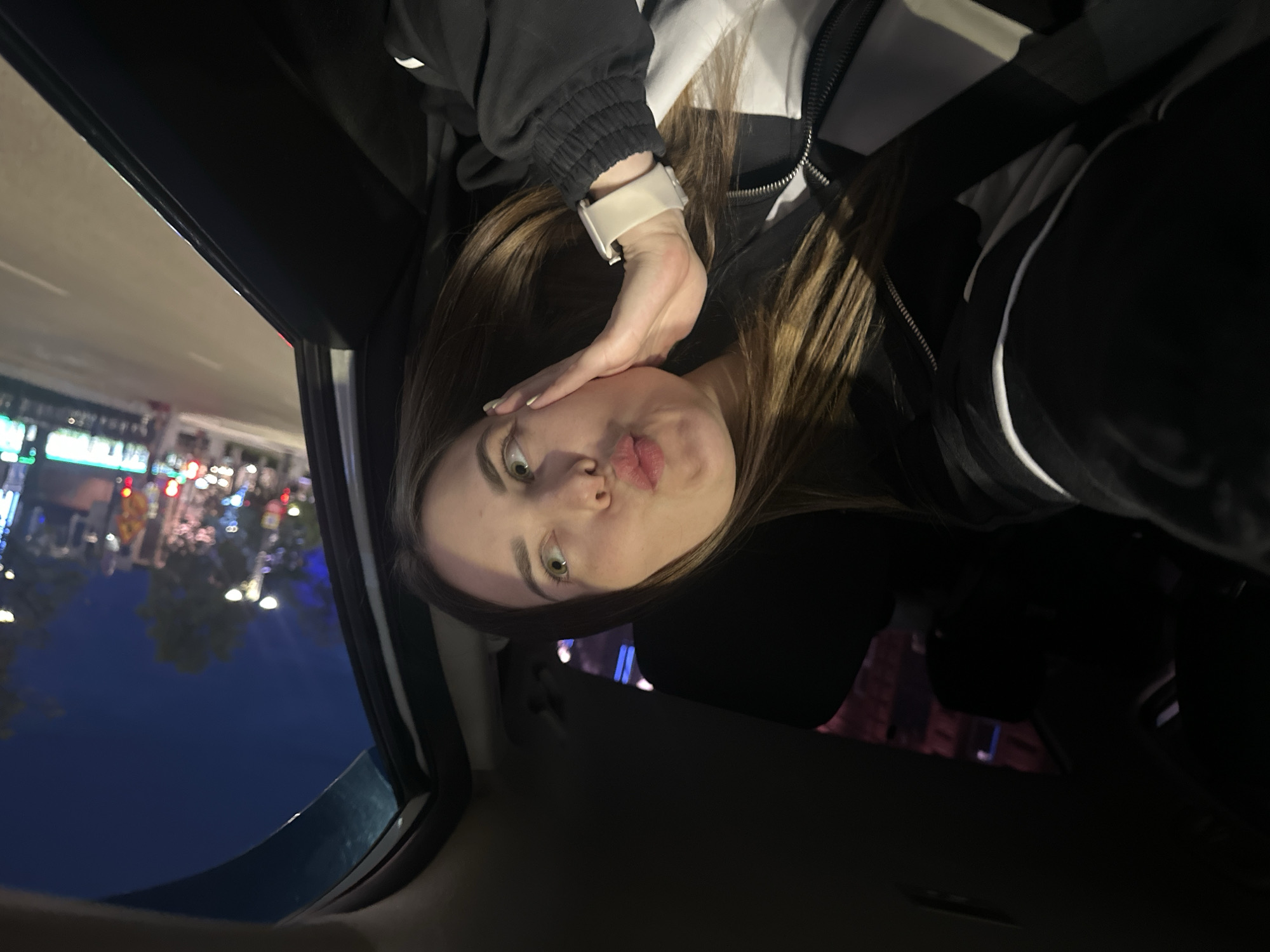 A young woman with long brown hair is sitting in the passenger seat of a car at night, taking a selfie. She is wearing a black and white jacket and a white watch. She is making a kissing face and resting her cheek on her hand. The car appears to be in motion, as evidenced by the blurred lights and scenery outside the window. The background shows a city street with buildings and streetlights. The sky is dark blue, indicating it is nighttime. The exact location in Perm, Russia, cannot be determined from the image.