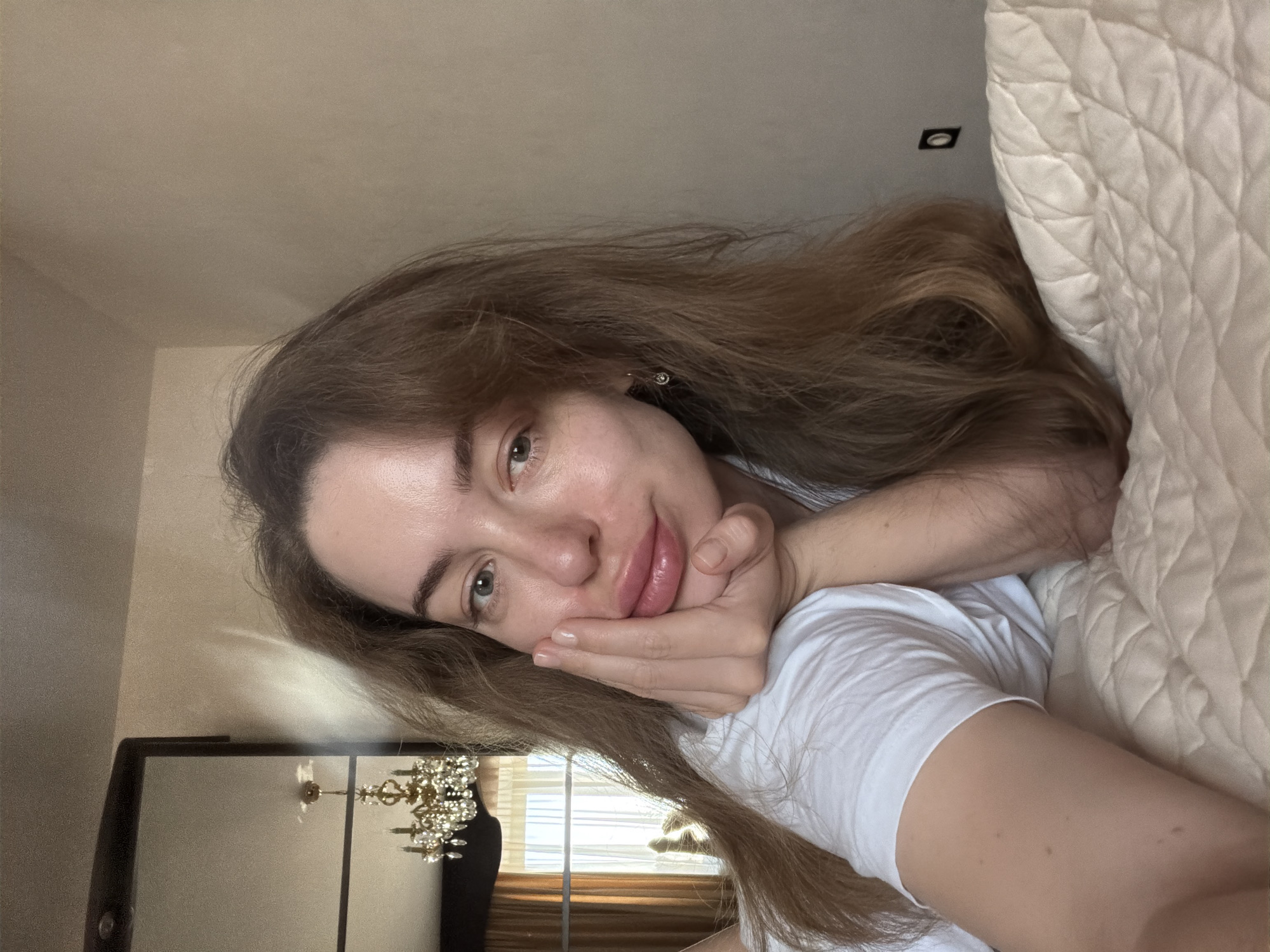 A female individual with light brown, wavy hair and light-colored eyes is depicted in a close-up selfie, lying prone or reclined on a quilted, light-beige bedding surface. She is wearing a white short-sleeved t-shirt. Her right hand, with fingers visible, supports her chin. A small, gold-toned hoop earring with a dangling jewel is present on her right earlobe. Her lips are full and exhibit a light pink tint.
The immediate background on the right is a plain, light-beige wall, featuring a dark rectangular electrical outlet or switch plate. To the left, a large dark-framed mirror reflects an ornate, multi-tiered crystal chandelier with gold-colored metalwork, which appears to be illuminated. Also visible in the mirror's reflection is a window draped with light-colored sheer curtains and heavier, gold-toned opaque drapes. Beyond the window, an exterior view reveals what appears to be another building structure.
The scene is consistent with an indoor residential setting within the city of Khimki, Russia, though specific street or address details are not discernible from the image.