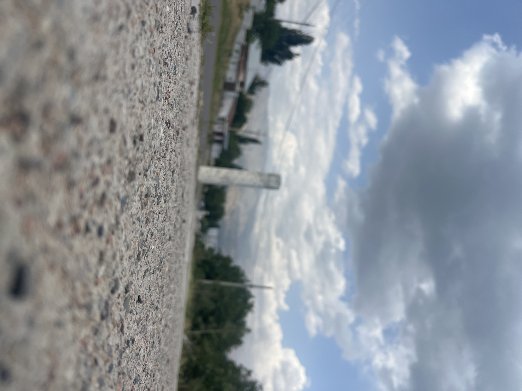 The image depicts a low-angle view of a gravelly road with a village scene in the background. The road surface is prominent in the foreground, showing small stones and pebbles. In the middle ground, a few houses with light-colored walls and darker roofs are visible, nestled among trees. A tall, slender white monument or signpost stands to the right of the houses. Beyond the village, hills are faintly discernible under a sky filled with a mix of white and dark, cloudy formations. Power lines are also visible crisscrossing the scene. The overall impression is of a rural or semi-rural setting, likely in Yusuphana, Uzbekistan, under a partly cloudy sky. There are no people or animals visible in the image.
