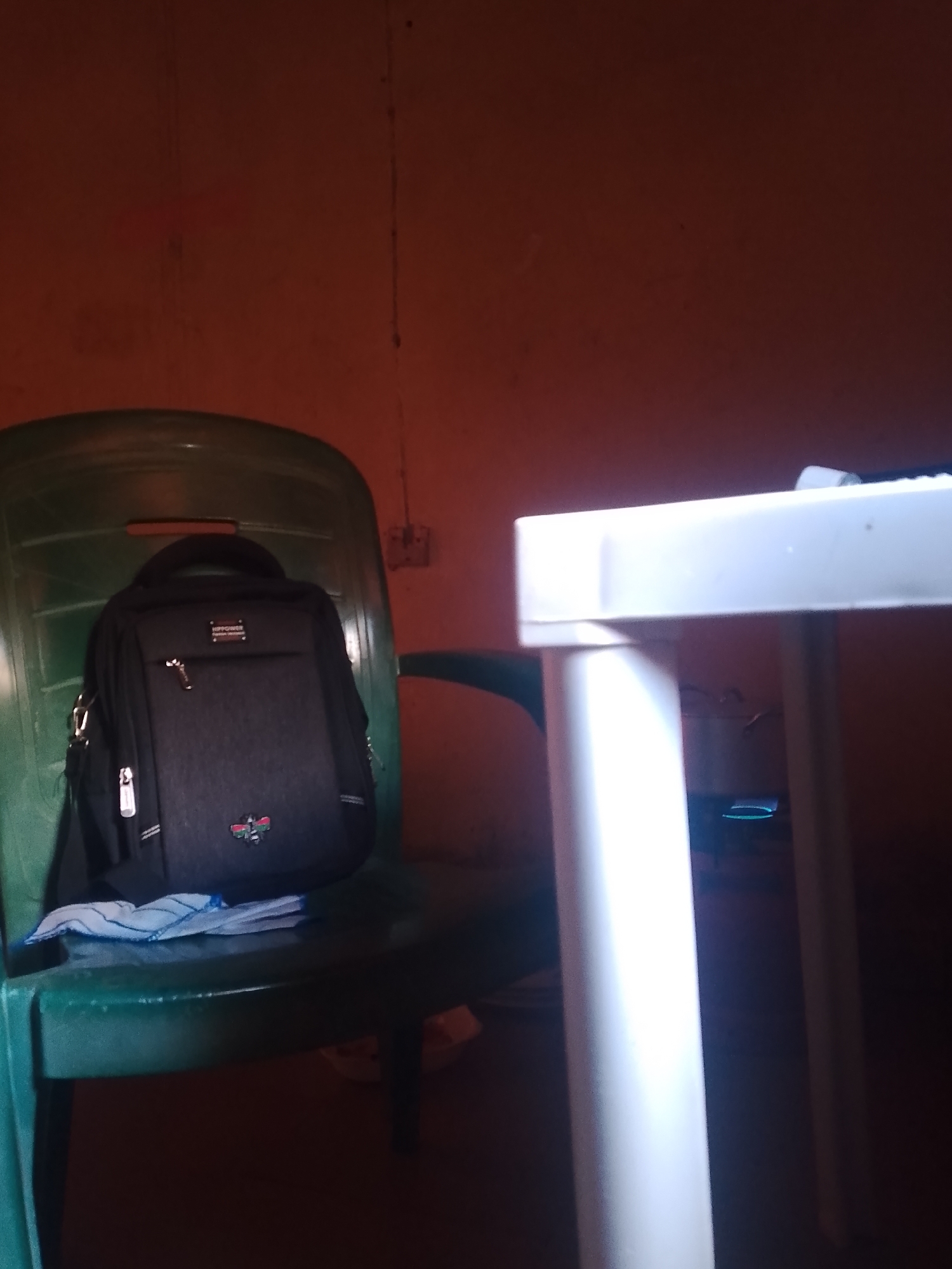 The image shows a black messenger bag placed on a green plastic chair. A blue and white cloth is draped over the seat of the chair, partially underneath the bag. To the right of the chair is a white table with visible legs. The background consists of an orange-brown wall. The lighting suggests it is likely indoors, possibly during daytime or with artificial lighting, casting shadows and creating a dim atmosphere. There are no people visible in the image. The scene is simple and domestic, with common household objects. There is no discernible activity taking place; the objects are stationary. A small tag on the bag has text that is too blurry to read clearly, but appears to have some lettering and possibly a logo. The overall impression is one of a quiet, perhaps informal setting within a room.