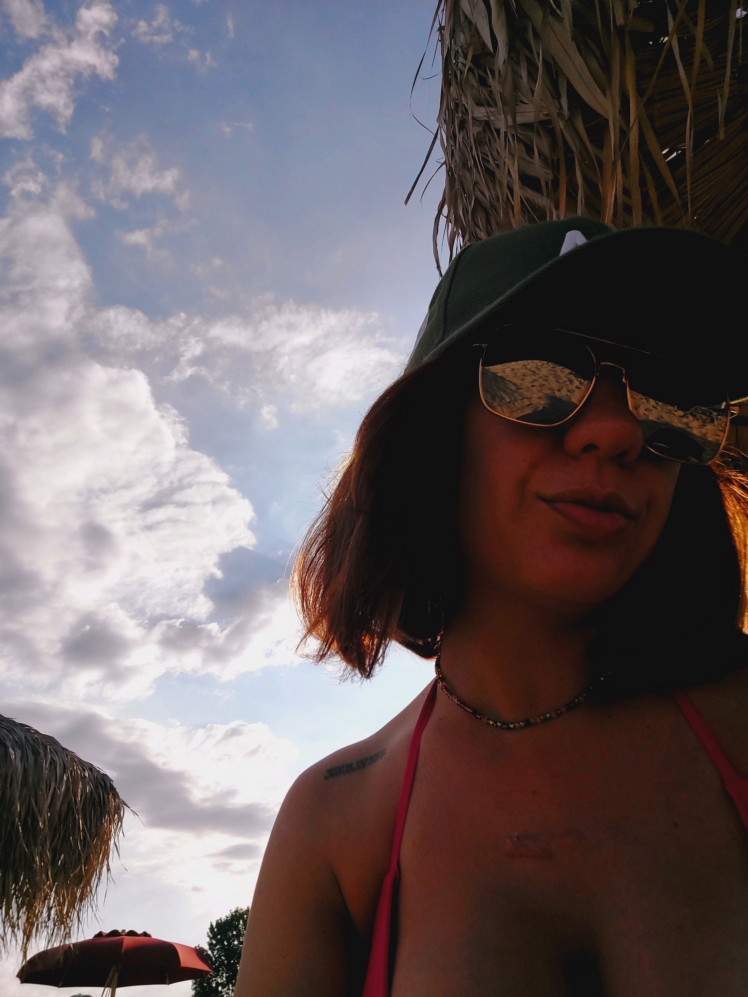 A woman, wearing a green baseball cap and reflective sunglasses, poses for a selfie in Sozopol, Bulgaria. She has brown hair and is wearing a pink bikini top and a beaded necklace. A tattoo is visible on her left shoulder blade and another small tattoo is on her chest. She is outdoors, with a beach umbrella and palm fronds visible in the background. The sky is partly cloudy.