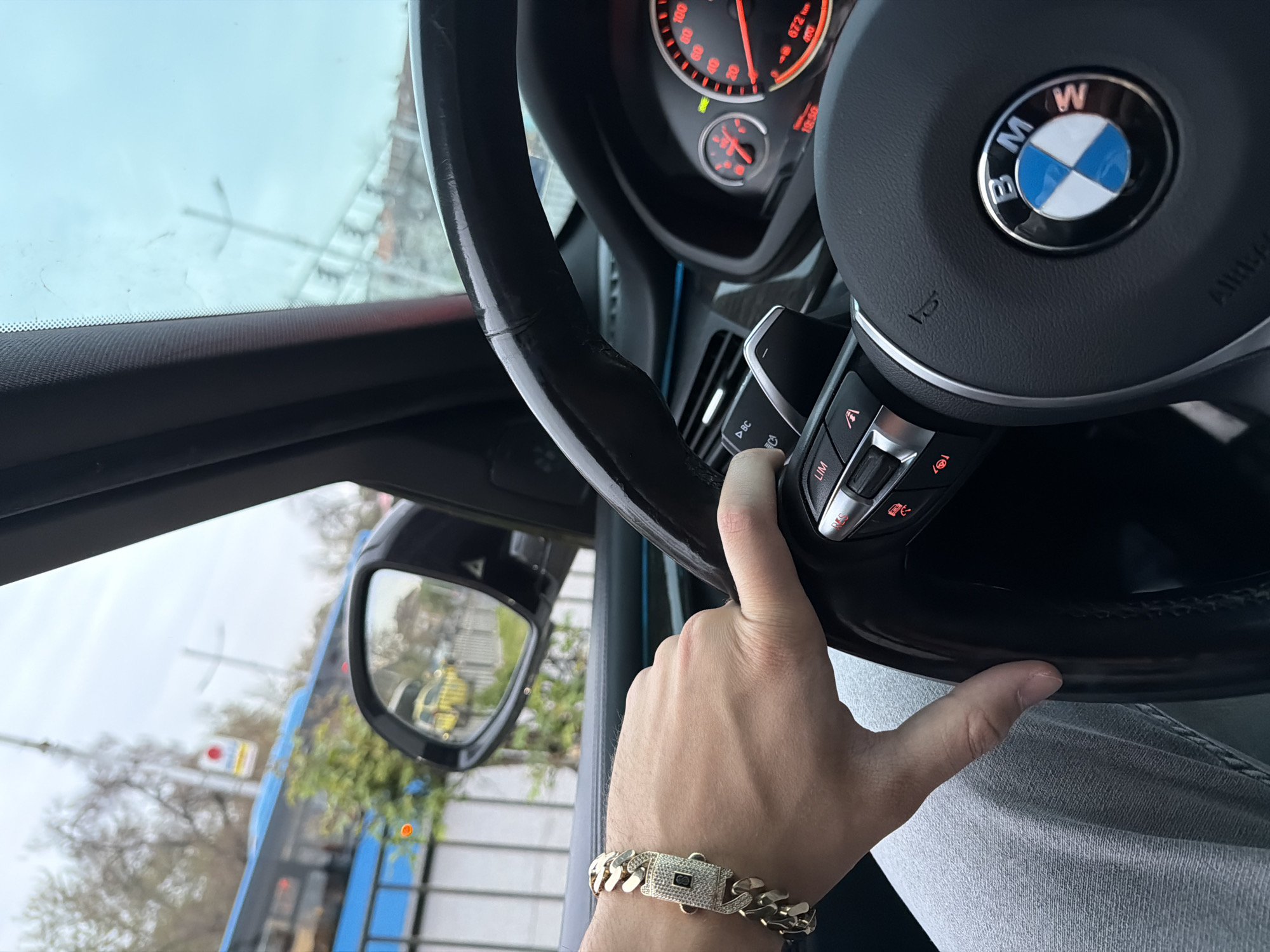 The image provides a close-up perspective from inside a car, focusing on a person's right hand gripping the steering wheel of a BMW vehicle.
The primary subject is a person's hand, adorned with a prominent gold-colored Cuban link bracelet featuring a rectangular clasp embellished with clear stones. The hand is positioned on the right side of the black leather steering wheel, with fingers near the control buttons.
The car's interior features a BMW steering wheel with the iconic blue and white logo at its center. Visible on the steering wheel are various control buttons, including those marked "LIM" (likely for speed limiter) and symbols for audio or cruise control. The dashboard instruments, partially visible behind the steering wheel, are illuminated in red and orange, showing a speedometer with numbers up to "100" and "120" (implied by "100"), and a tachometer. A digital display reads "19:50," indicating the time, and "672 k," likely a distance reading in kilometers.
Through the car's windshield and side mirror, an exterior scene is visible. The sky appears overcast and bright. The windshield shows some streaks, possibly from dirt or rain. The side mirror reflects blurred shapes, including what seems to be a yellow object and parts of a building or structure. Faintly visible through the windshield are blurred trees and some distant architectural elements.
The activity depicted is a person either driving or paused in a car, with their hand on the steering wheel, suggesting readiness to operate the vehicle. The time of day is 7:50 PM as indicated by the dashboard clock. There are no clear location cues beyond what appears to be a city or suburban street. No emotions are discernible.