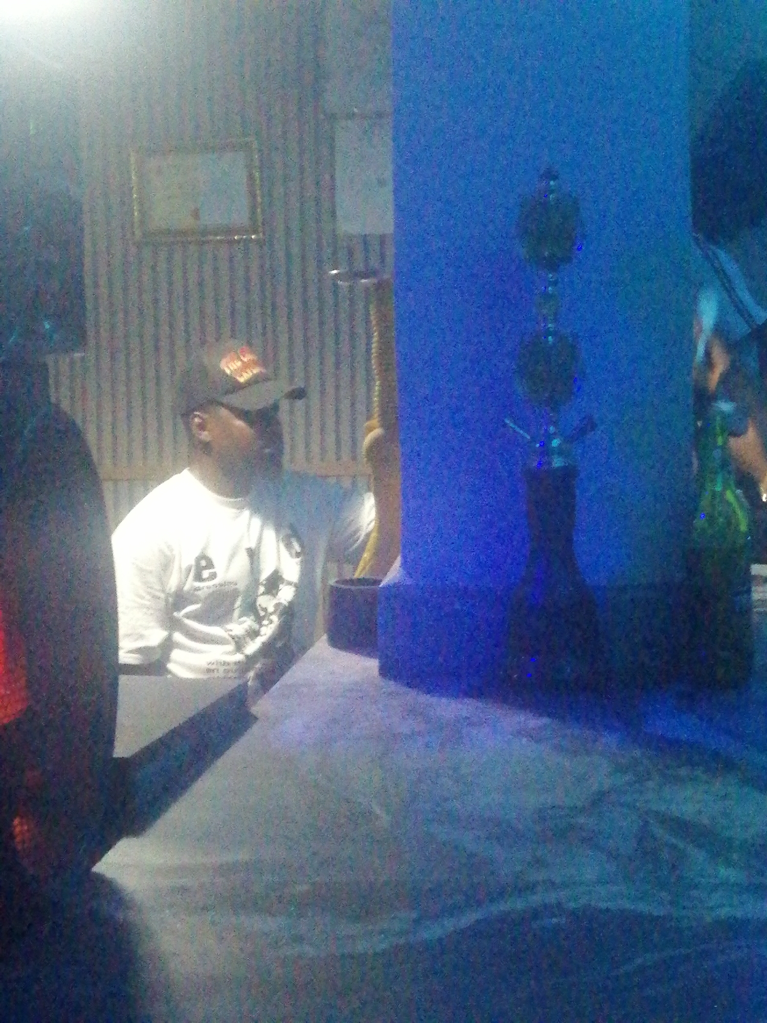 The image captures an indoor scene, likely a lounge or bar in Jos, Nigeria, characterized by dim lighting and a social atmosphere. The primary subject is a man seated at a table, viewed from the chest up. He is wearing a white t-shirt with a logo and text, and a dark baseball cap with red lettering that appears to say "The CITI" or similar. The man is looking towards the right side of the frame, his expression not clearly visible due to the low light and angle.
The setting includes a patterned wall with vertical stripes in the background to the left, featuring a framed picture or mirror. To the right, a solid blue wall or partition is visible. A dark-colored table, possibly covered with a cloth, occupies the foreground. On the table, two hookahs are prominent: a dark-colored one in the immediate foreground and a slightly lighter one further back, both against the blue background. A green bottle is also visible on the table behind the hookahs. The low light conditions give the image a grainy texture, suggesting it was taken in the evening or night. The overall impression is that of a relaxed social environment.