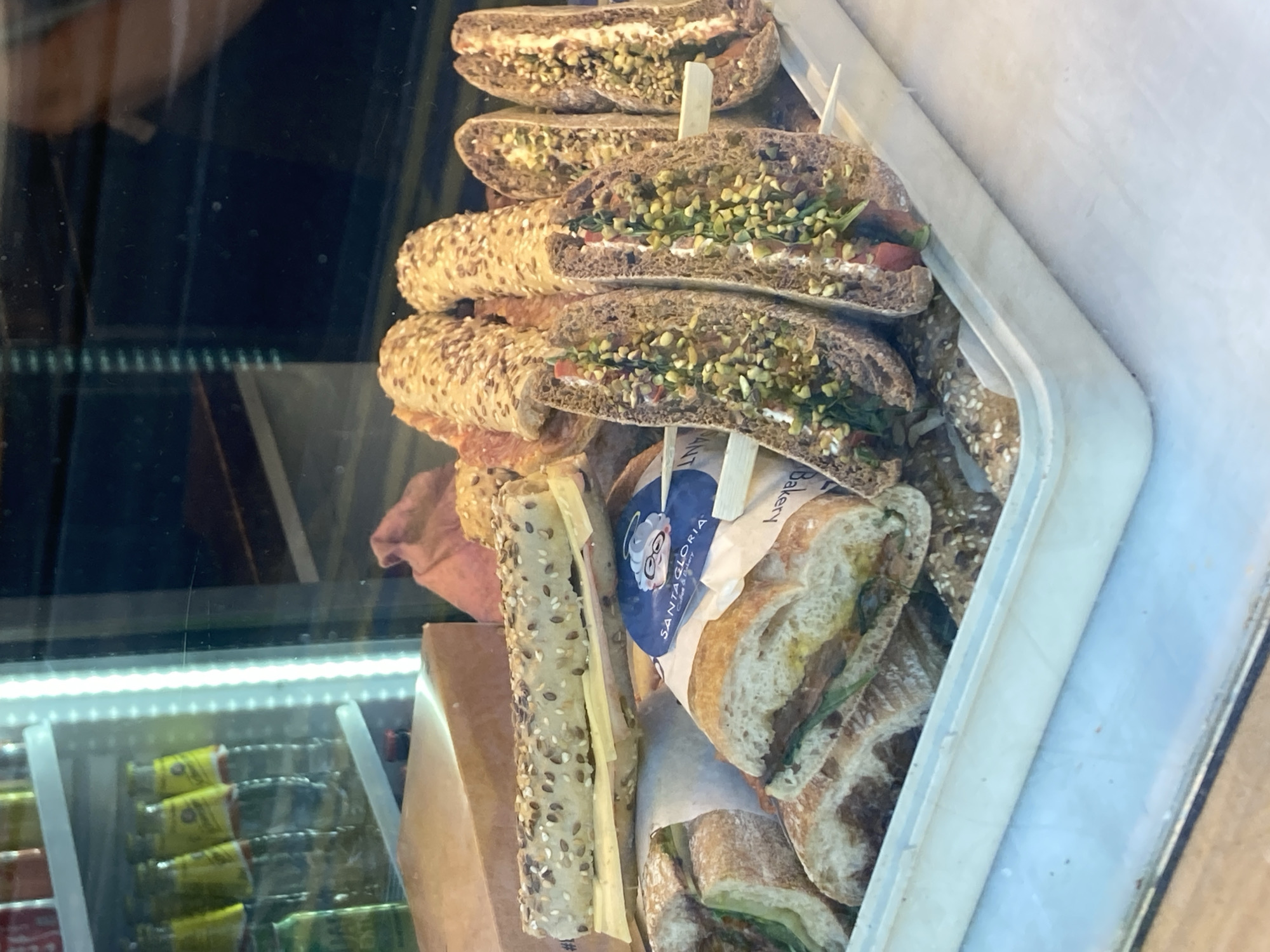 The image shows a close-up, high-angle view of several sandwiches displayed in a refrigerated glass case, likely in a bakery or deli. The sandwiches are made with dark, seeded bread and filled with what appears to be cream cheese, chopped green herbs, and possibly other vegetables or nuts. Some sandwiches are cut in half, revealing their fillings, while others are whole. One sandwich is partially wrapped in paper with the "SANTA GLORIA" logo, featuring a cartoonish elderly woman with glasses.
The setting is a commercial establishment, with a bright LED light strip visible along the bottom of the case, illuminating the products. Below the sandwiches, stacks of individually wrapped items, possibly snacks or drinks, are visible in another section of the refrigerated unit, some with yellow and green packaging. The overall impression is of a food display showcasing fresh, prepared sandwiches. The image is sharp and well-lit, highlighting the textures and colors of the food. The location context provided is Barcelona, Spain, which might suggest a local bakery or food chain. There are no people or overt indications of the time of day or weather. The focus is entirely on the food items.