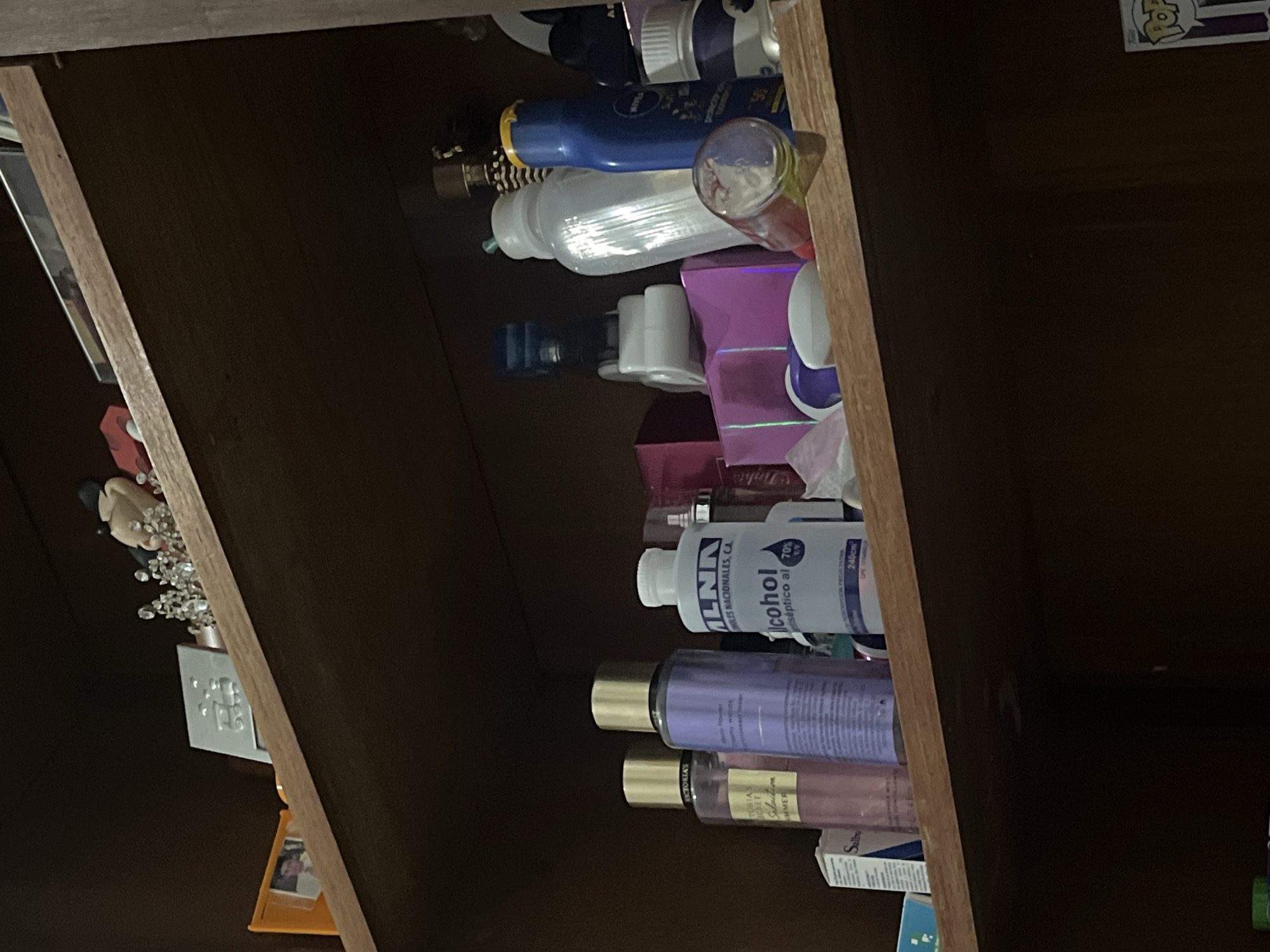 The image shows a cluttered shelf in a dimly lit room, likely in Santa Ana, Venezuela. The shelf is made of dark wood and is filled with various personal care products and decorative items.
On the top shelf, a small framed photo of a man is visible in an orange frame, alongside decorative trinkets resembling crowns and figurines. There is also a small red toy car.
The second shelf, which is the primary focus, contains several bottles of lotions and perfumes, including two bottles from Victoria's Secret, one pink and one purple. A bottle of antiseptic alcohol with a white cap is also present. Other items include pink and purple boxes, a clear plastic bottle, a blue bottle of Nivea sunscreen, and a white bottle with a purple cap. A small glass container lies on its side, showing colorful contents.
The bottom shelf is largely obscured by shadows, but some items are vaguely visible, including what appears to be a Funko Pop figure on the right side. The overall impression is one of a personal space that is not meticulously organized.
