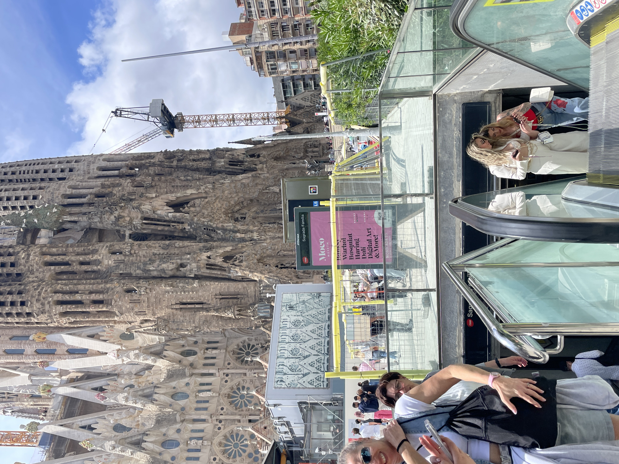 This image shows the Sagrada Familia basilica in Barcelona, Spain. The basilica is a large, intricately detailed stone building. Construction cranes are visible near the top of the building. In the foreground is the entrance to the Sagrada Familia metro station, with signs advertising the Moco Museum. There are people on the escalators going into the metro. Other people are visible on the street level near the entrance. The sky is blue with scattered clouds.