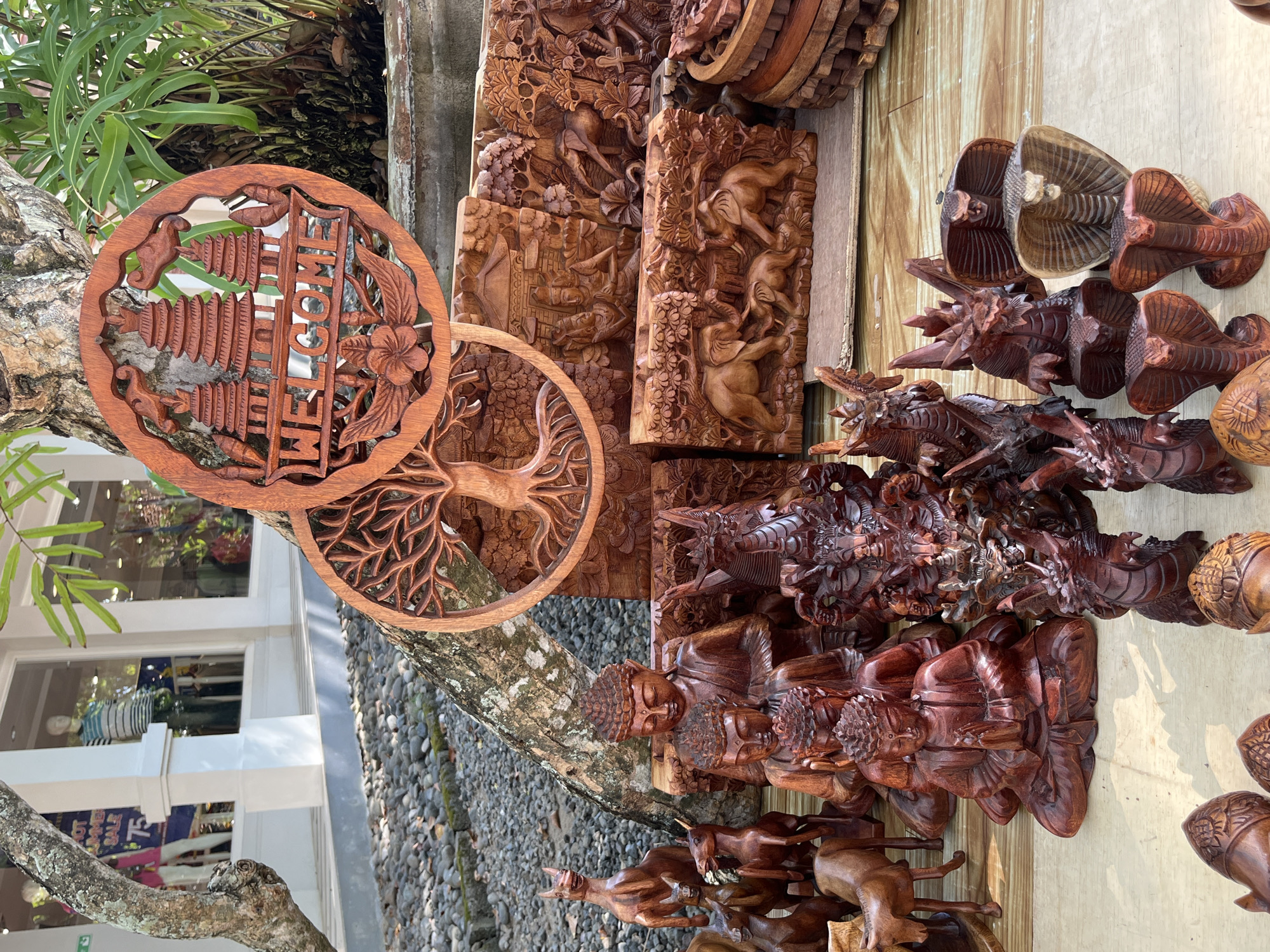 The image displays a collection of intricately carved wooden items for sale at an outdoor market stall. In the foreground, several wooden cobra figures are displayed prominently, some with flared hoods. To the left of the cobras are several dragon figures, also carved from wood. Behind these, a row of wooden Buddha statues are arranged, some in meditation poses. Further back, larger wooden carvings depict scenes with elephants and human figures, as well as a carved tree of life and a circular "Welcome" sign with temple motifs. The stall is set up on a wooden platform, with a backdrop of a tree and lush green plants. In the background, a building with large windows and mannequins visible inside suggests the presence of shops, characteristic of a commercial area. The ground is covered with small stones. Based on the style of the carvings and the general ambiance, the location is likely a tourist market in Kuta Selatan, Indonesia, known for its artisanal crafts.